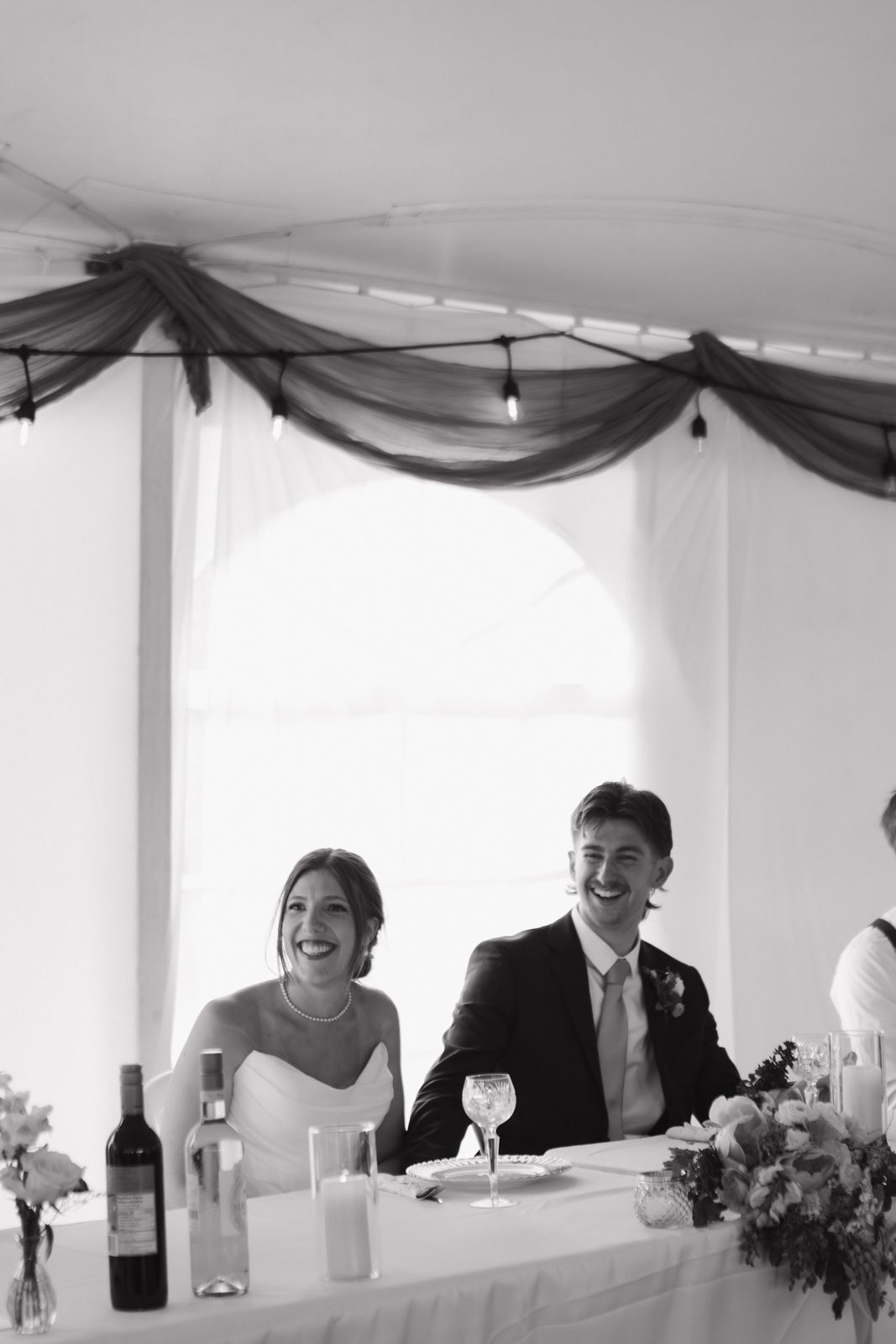 Black and white image of a bride and groom sitting at the head table listening to speeches.