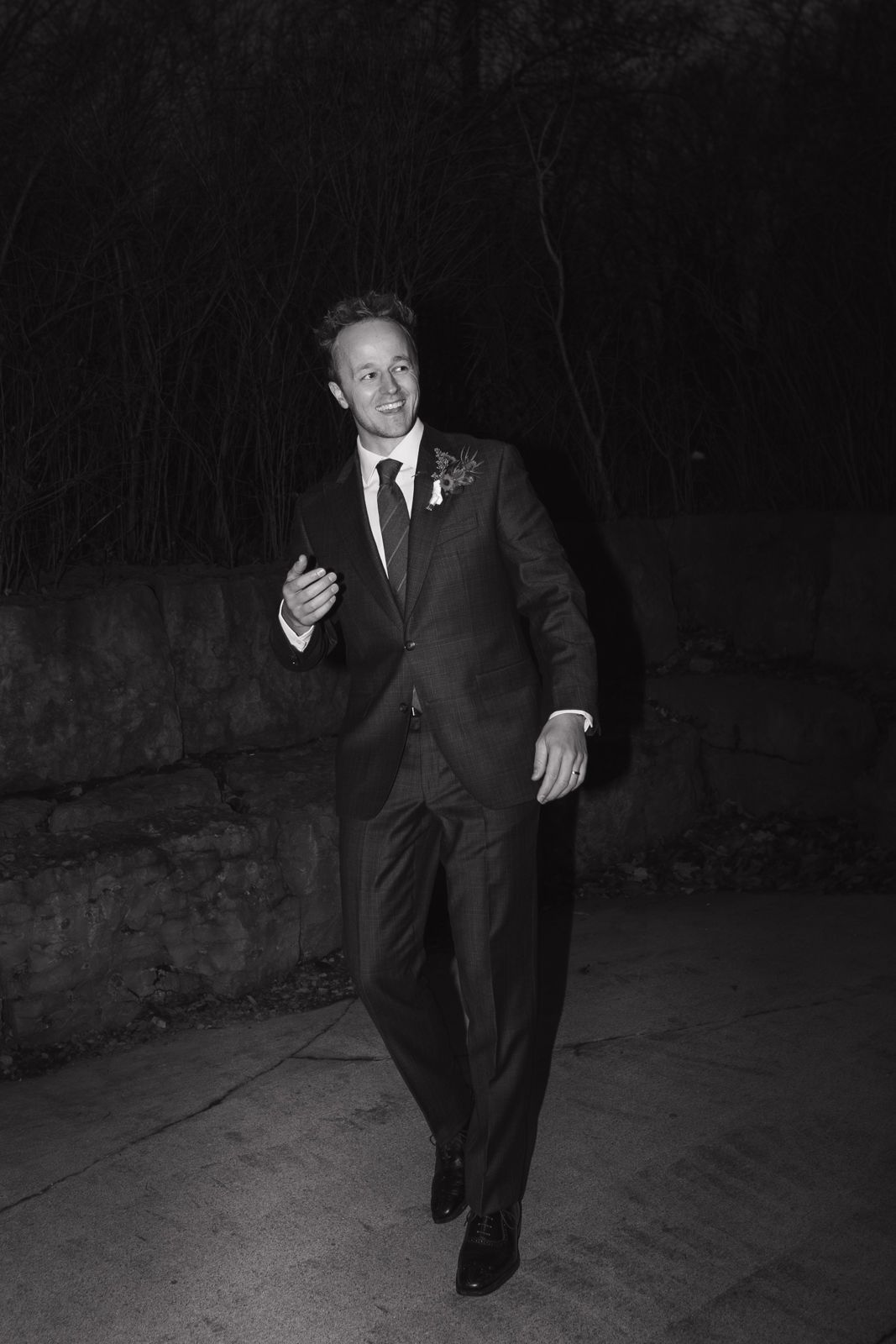 Black and white paparazzi-style image of a groom walking toward the camera. He is looking slightly to the right off-camera.