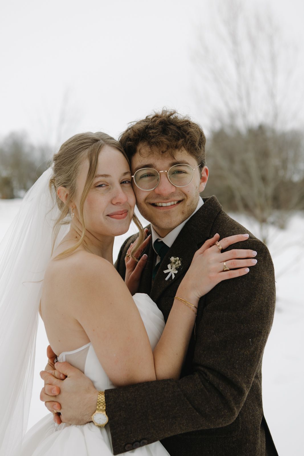 A bride and groom embracing and smiling toward the camera. They are outside in the snow.