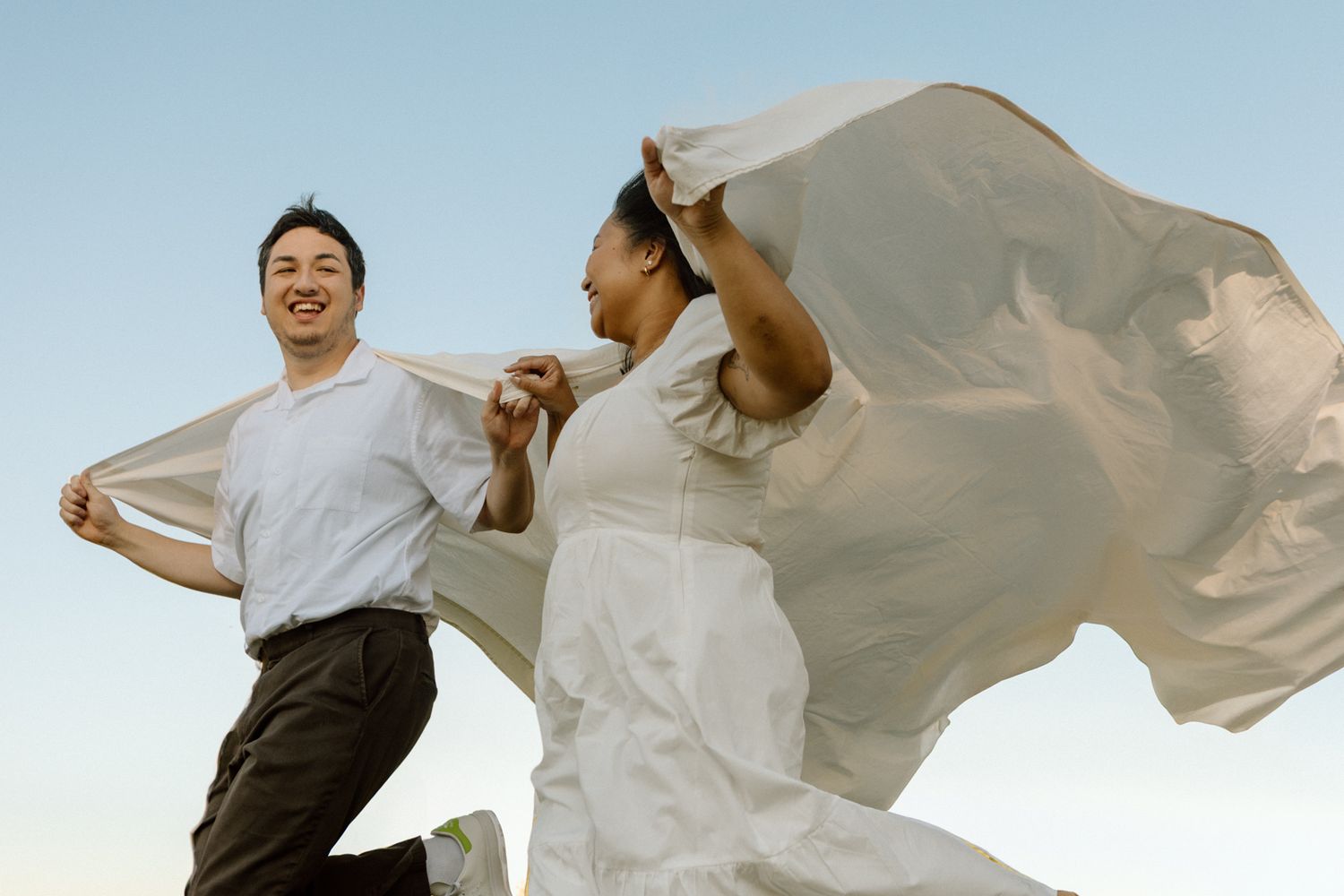 Couple running through a field holding a blanket caught in the wind, which trails behind them like a cape.