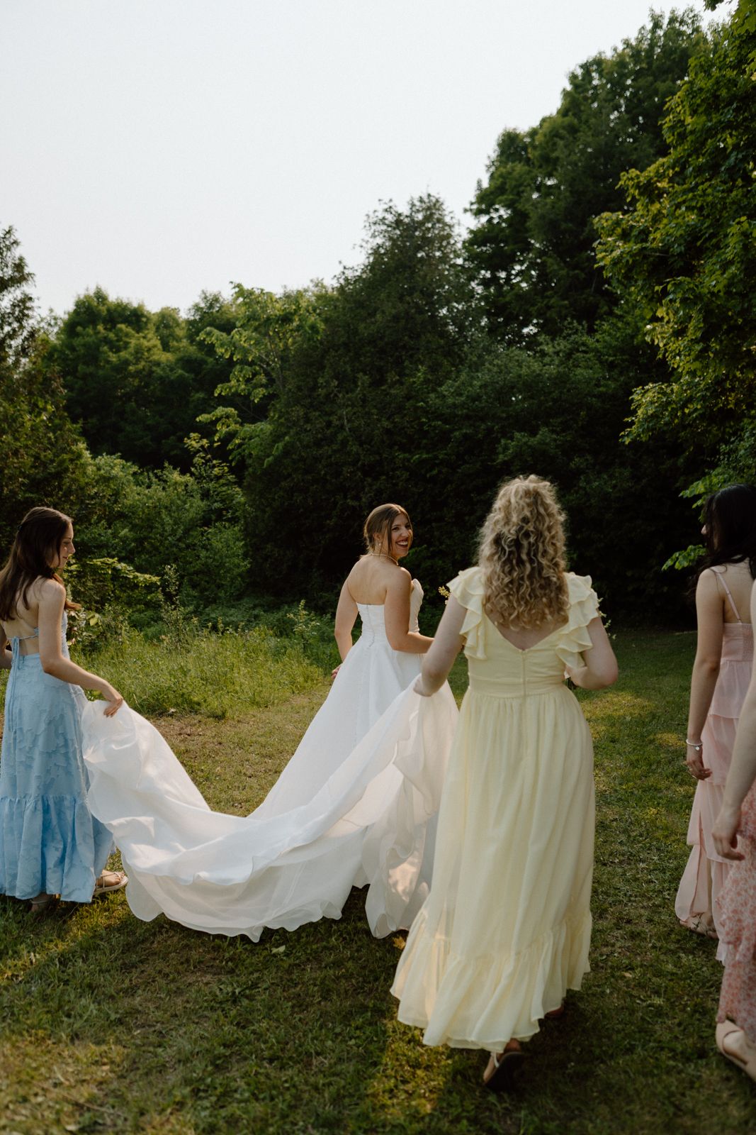 Bridesmaids in colourful mismatched dressed help a bride carry her train while she looks back at the camera and smiles.
