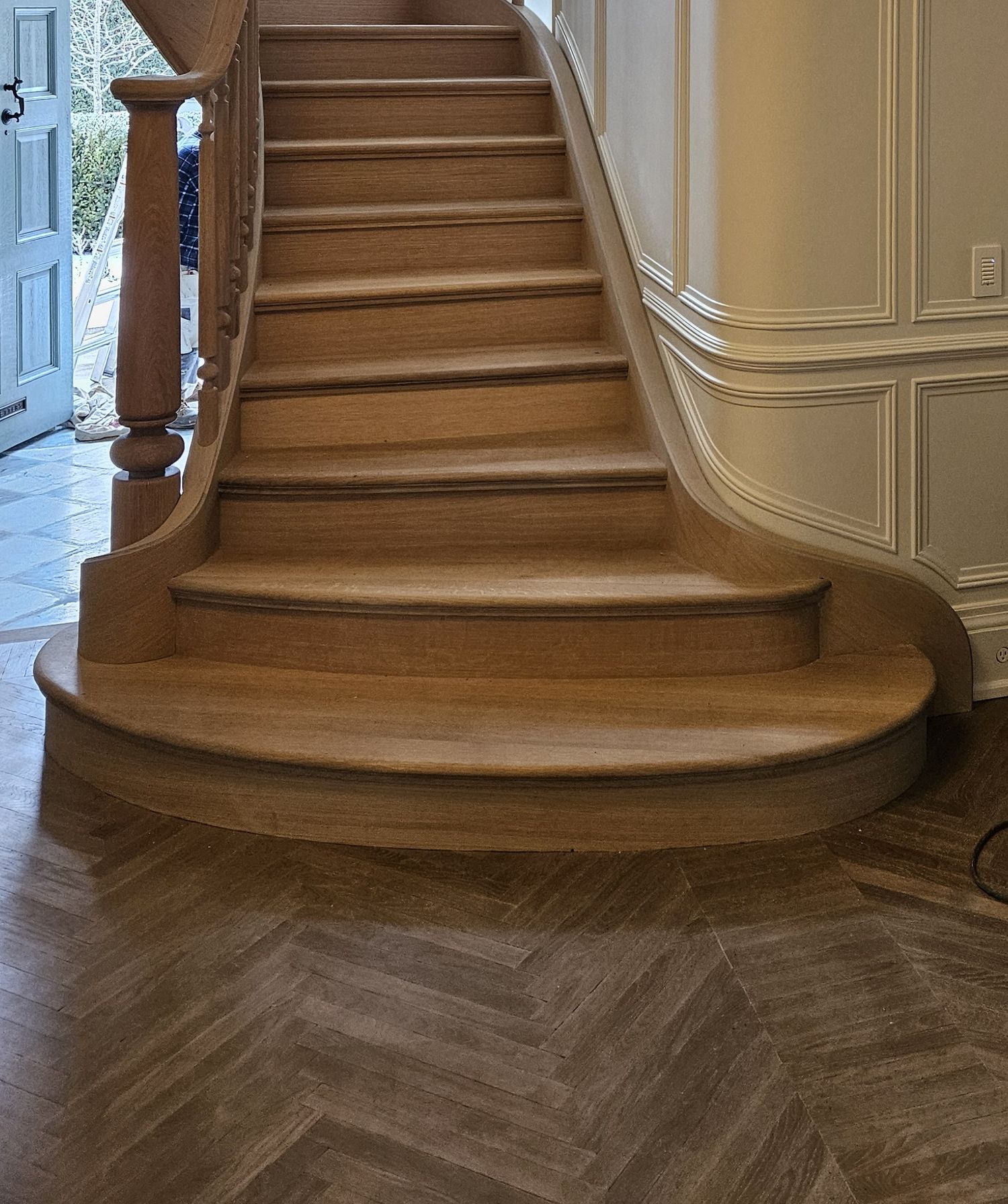 Elegant curved oak staircase with bullnose bottom steps and herringbone wood floor in upscale foyer.