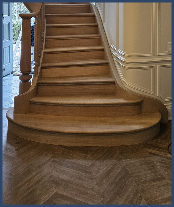 Stunning curved wood staircase with rich oak finish above a classic herringbone parquet floor.