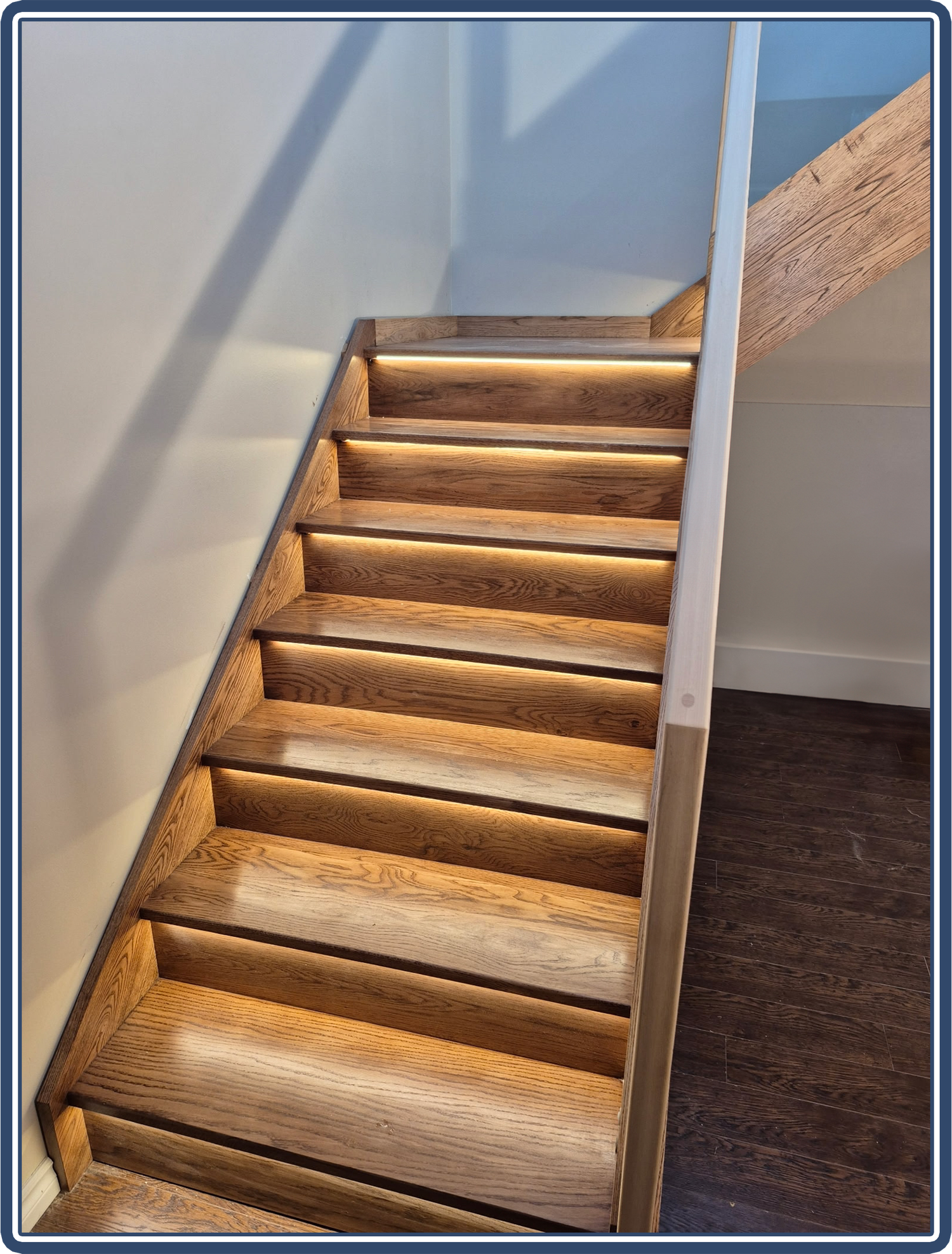 Oak staircase with LED under-step lighting and glass railing with a wooden handrail.