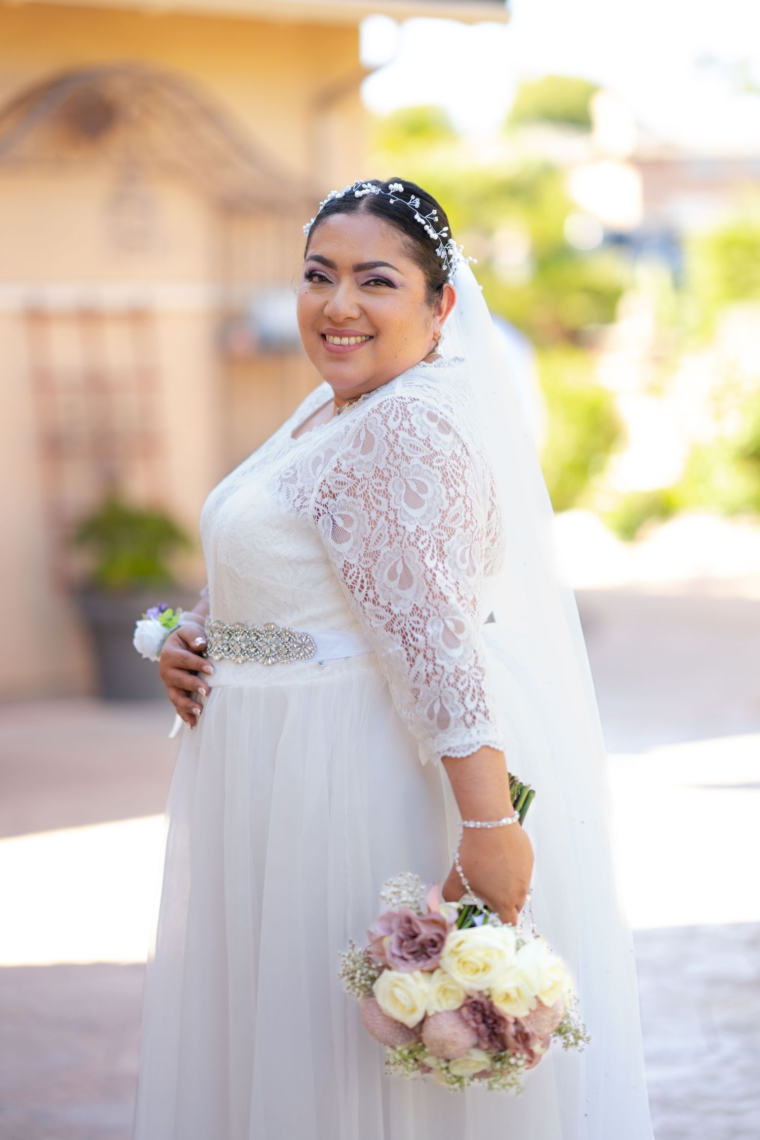 A bride in a white lace wedding dress with sleeves poses outdoors holding a pastel floral bouquet.