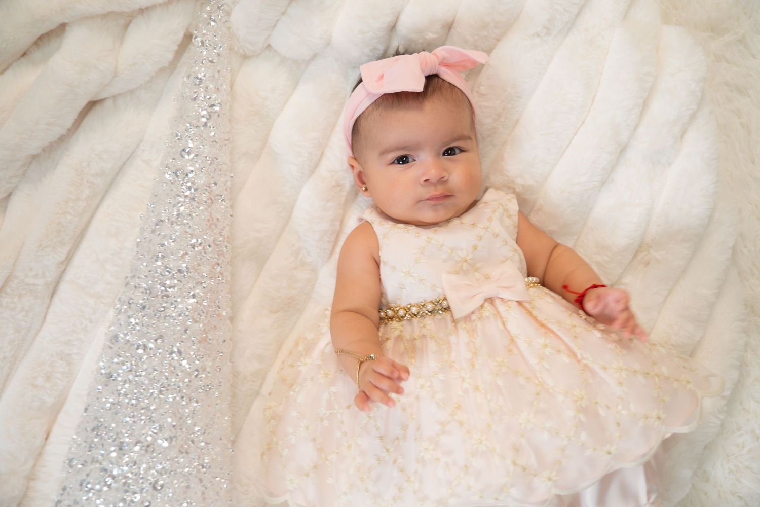 Baby wearing a pink lace dress and bow headband lies on white tulle fabric with sparkly glitter details.