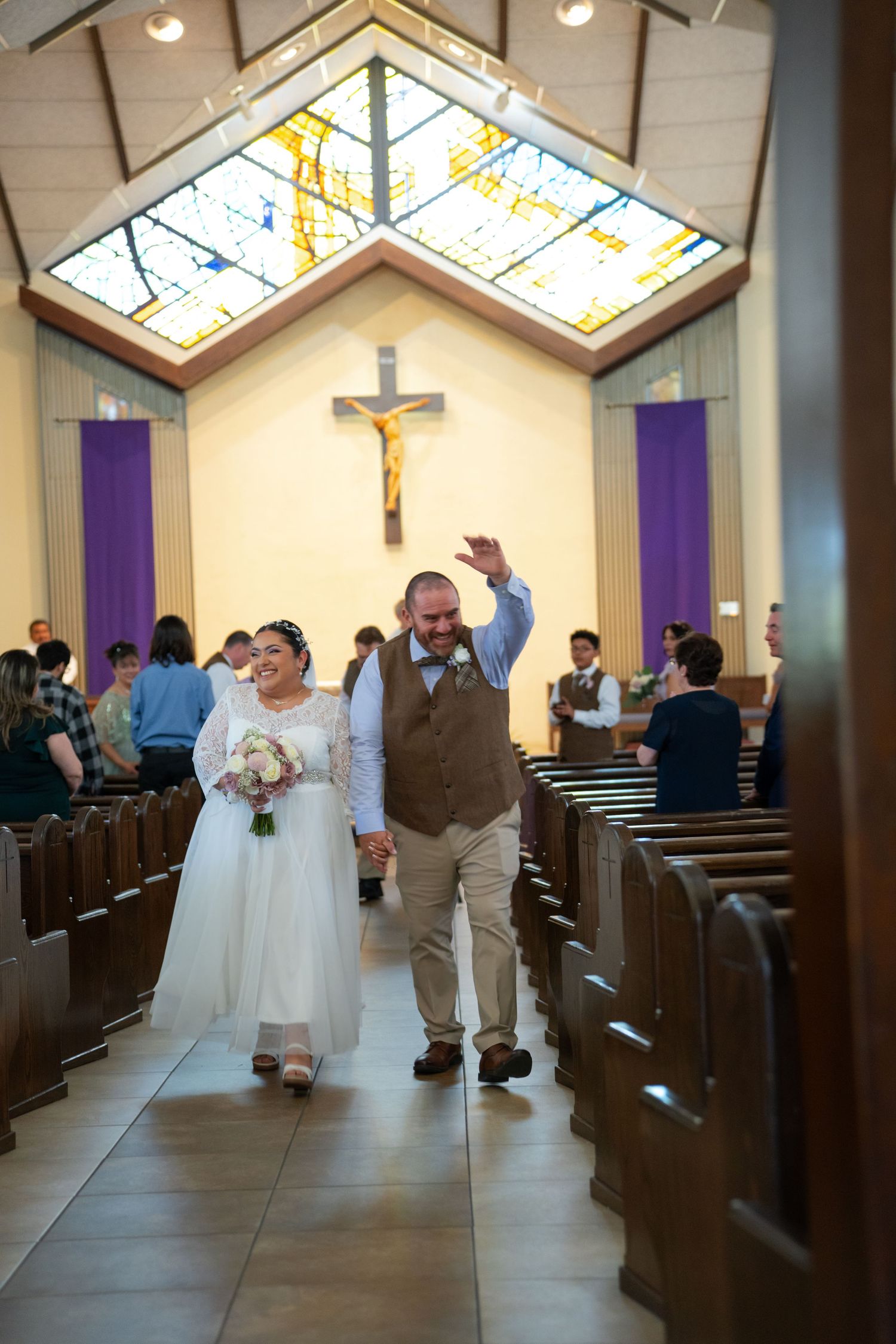 Newlyweds walk down the church aisle after their ceremony beneath stained glass windows and a crucifix.