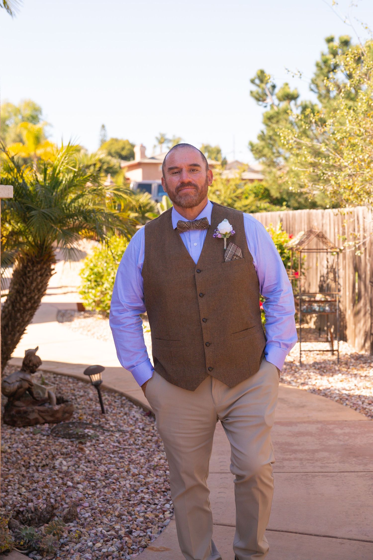 A groom in a brown vest and blue bowtie poses in a sunny garden setting with palm trees.