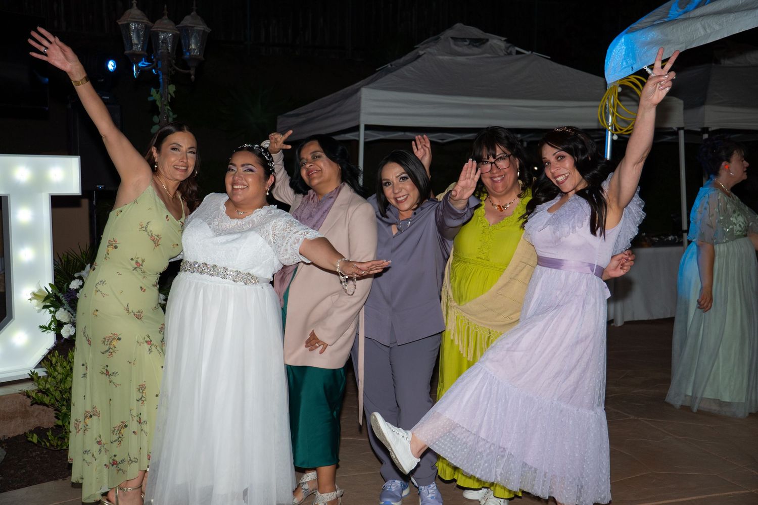 A group of people dancing and celebrating together at an outdoor evening reception under a white tent.