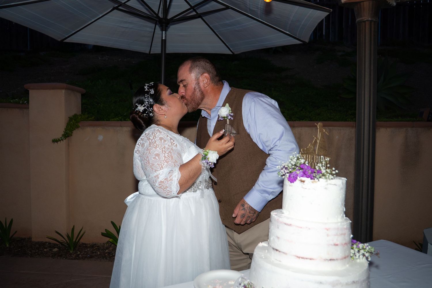 A romantic couple shares a kiss during their wedding reception near a white tiered cake with purple floral accents.