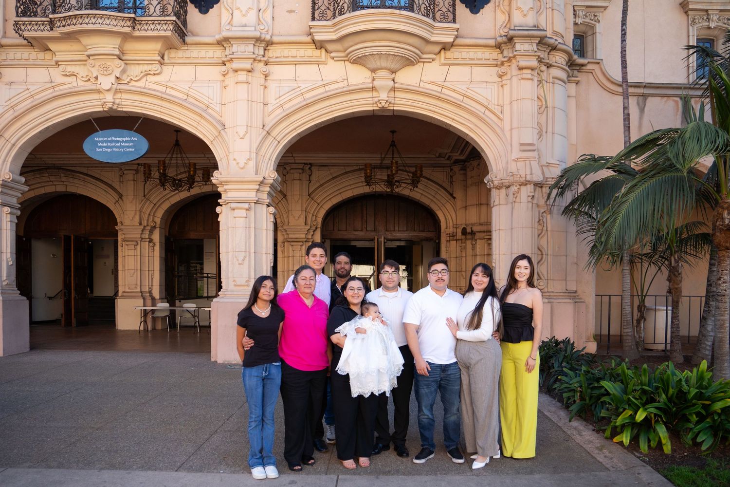 Group of people gathered in front of an ornate stone archway with Mediterranean-style architecture and palm trees.