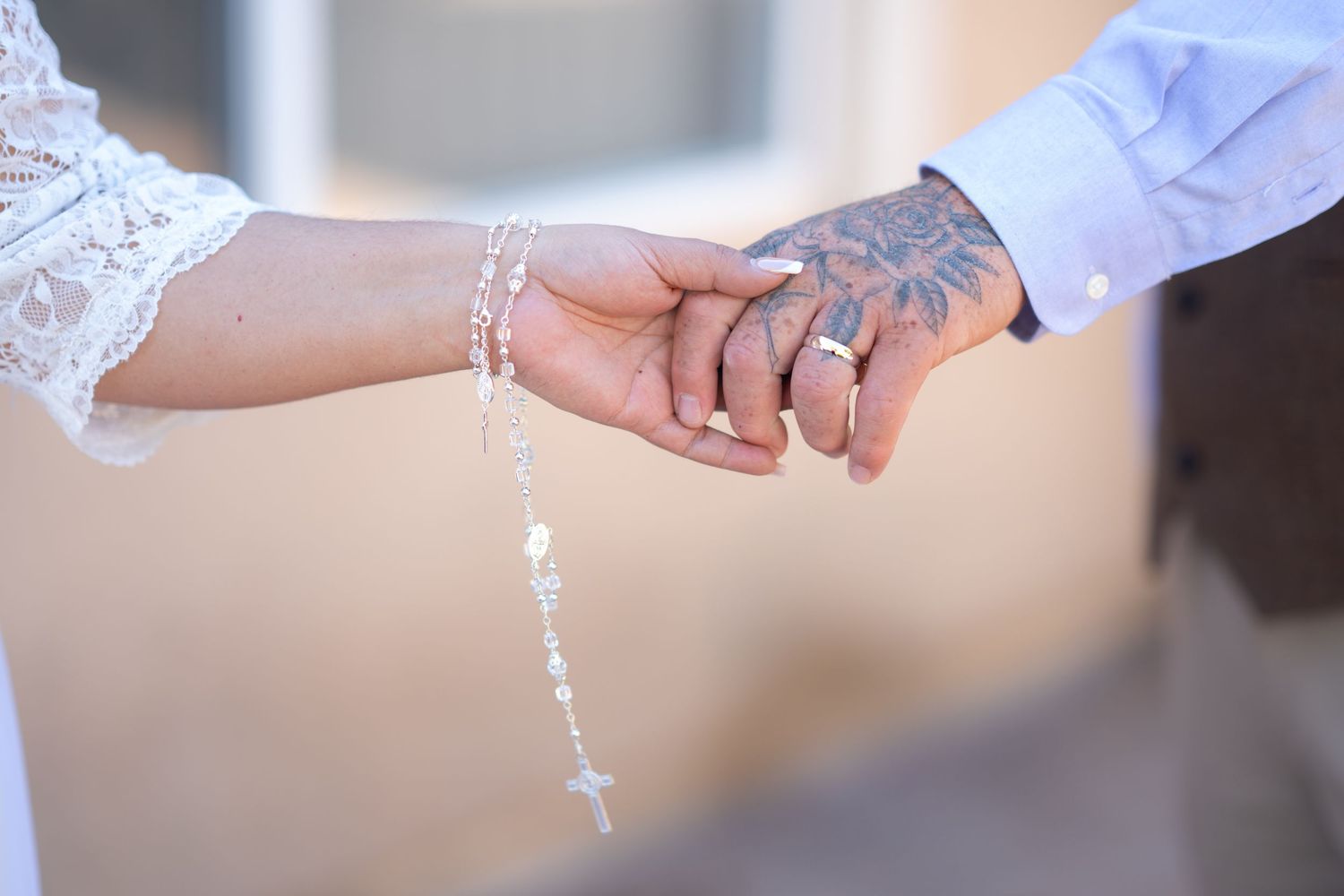 Close-up details of wedding rings and hands with delicate lace and rosary beads against a soft blurred background.