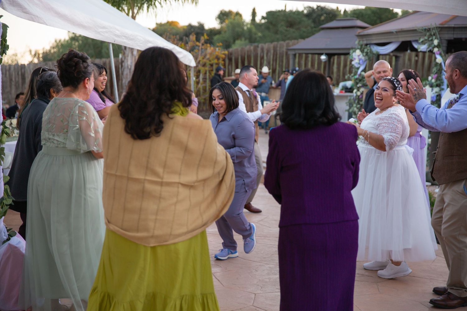 Guests in colorful formal attire gather at an outdoor wedding reception under white canopy tents.