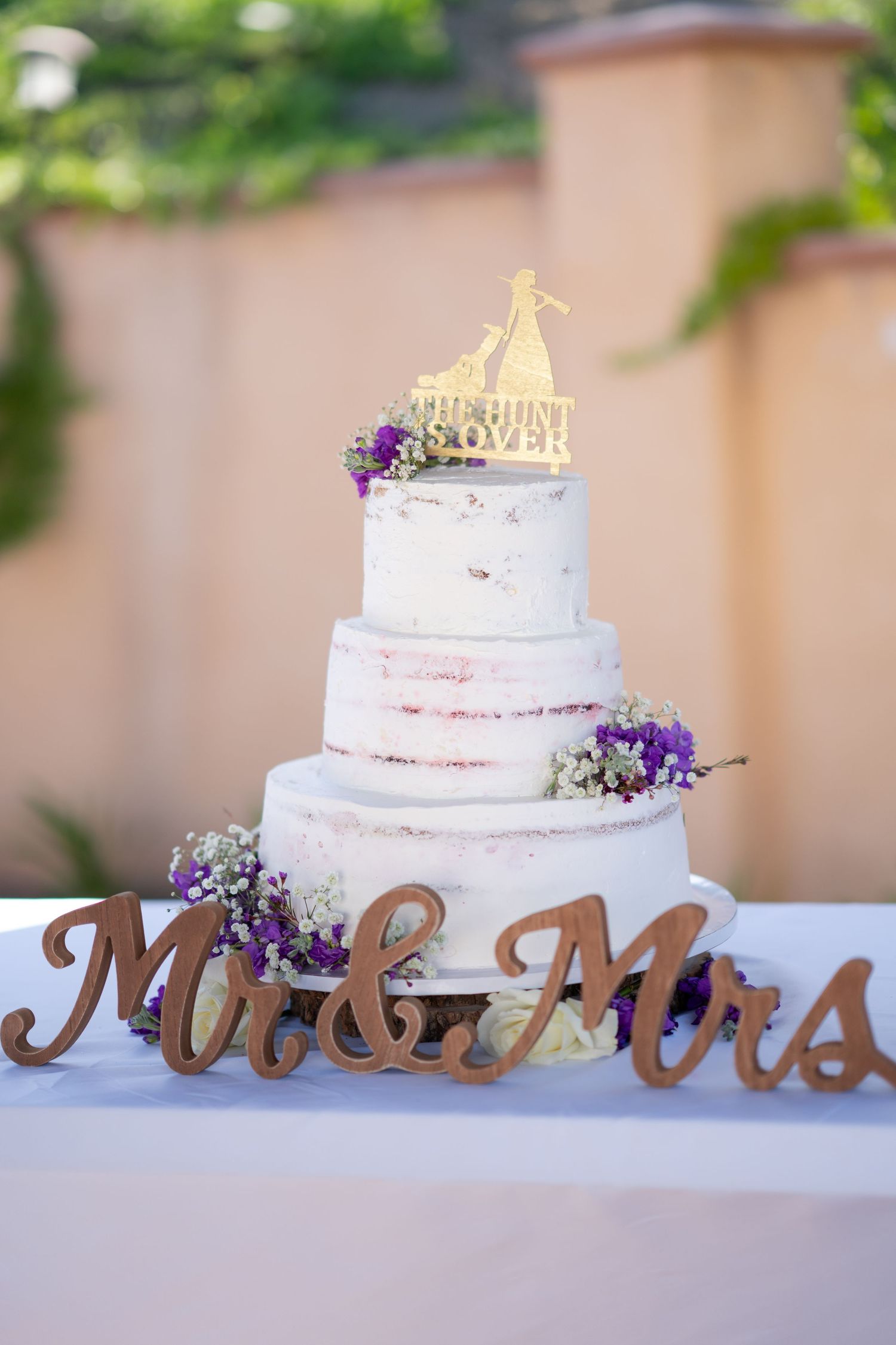 Elegant white wedding cake display with wooden Mr and Mrs sign and purple floral accents.