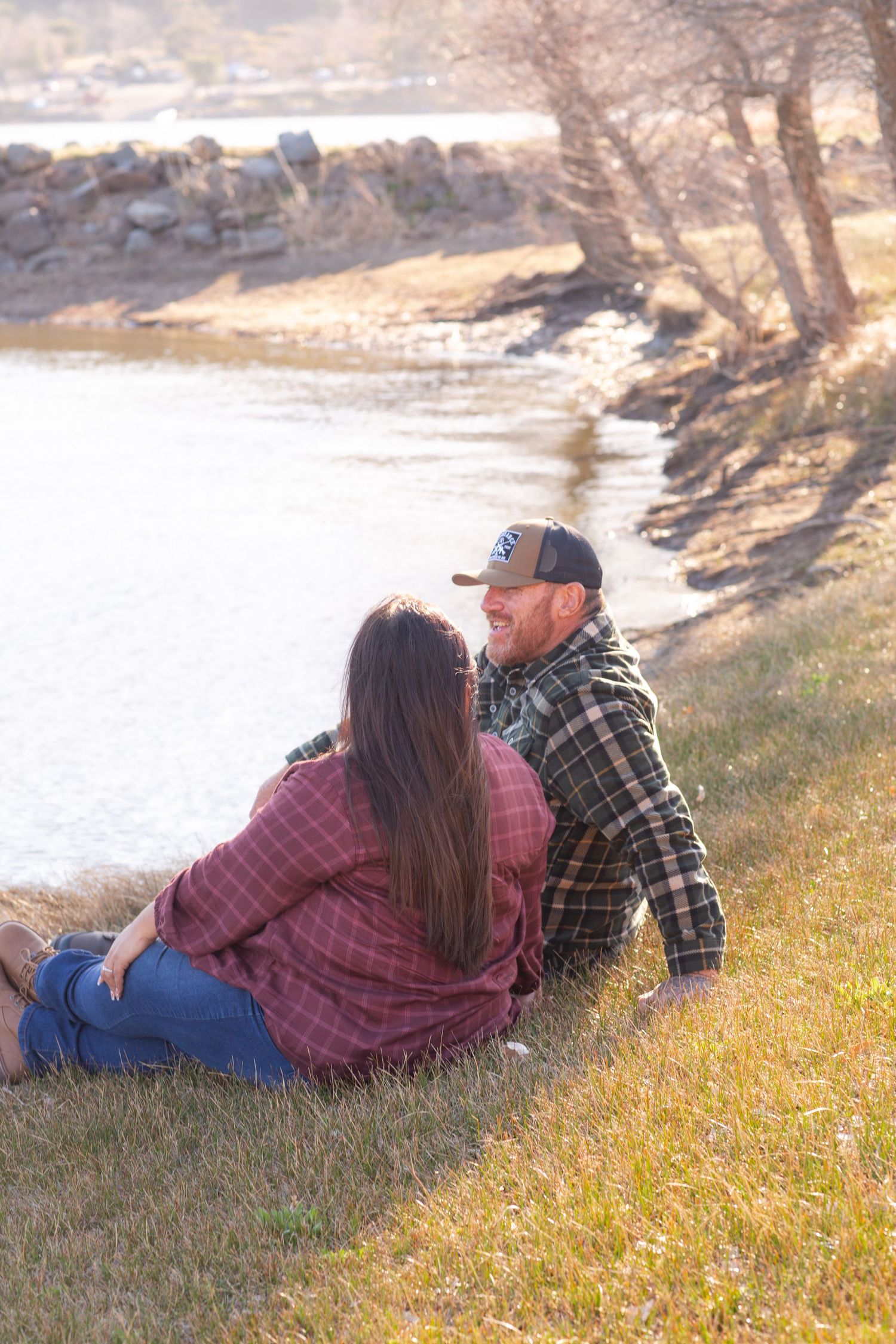 A couple in casual attire relaxes on a grassy hillside during golden hour near a lake.