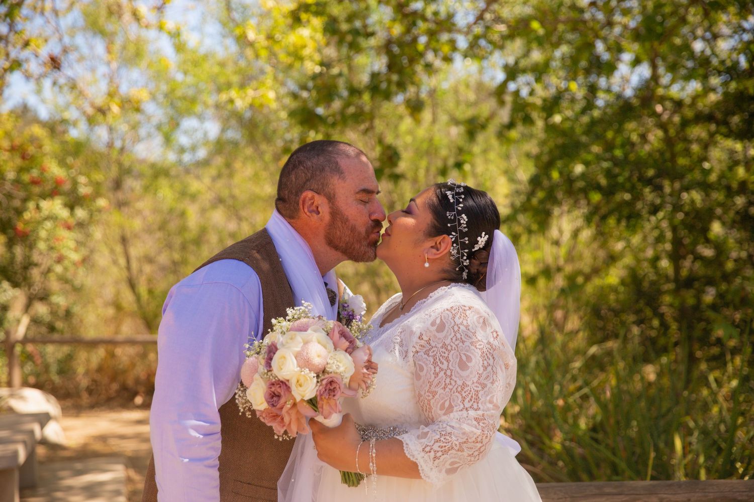 A romantic outdoor wedding moment with a couple kissing while holding a blush pink and white floral bouquet.