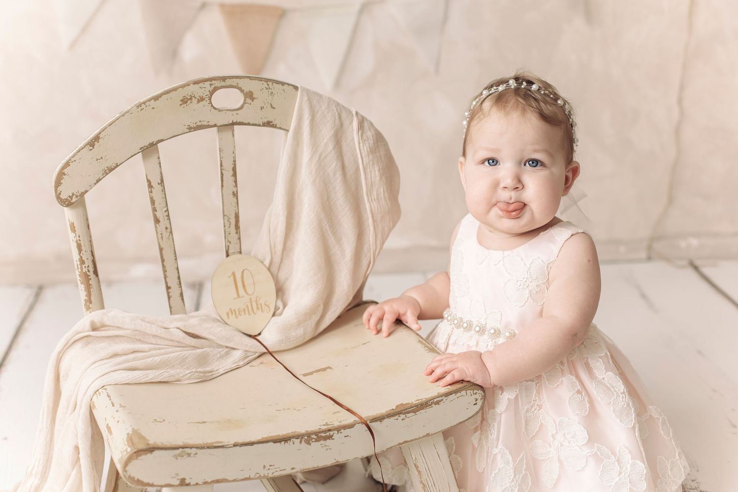 Adorable baby girl in white lace dress with floral headband posing beside a vintage distressed wooden chair.