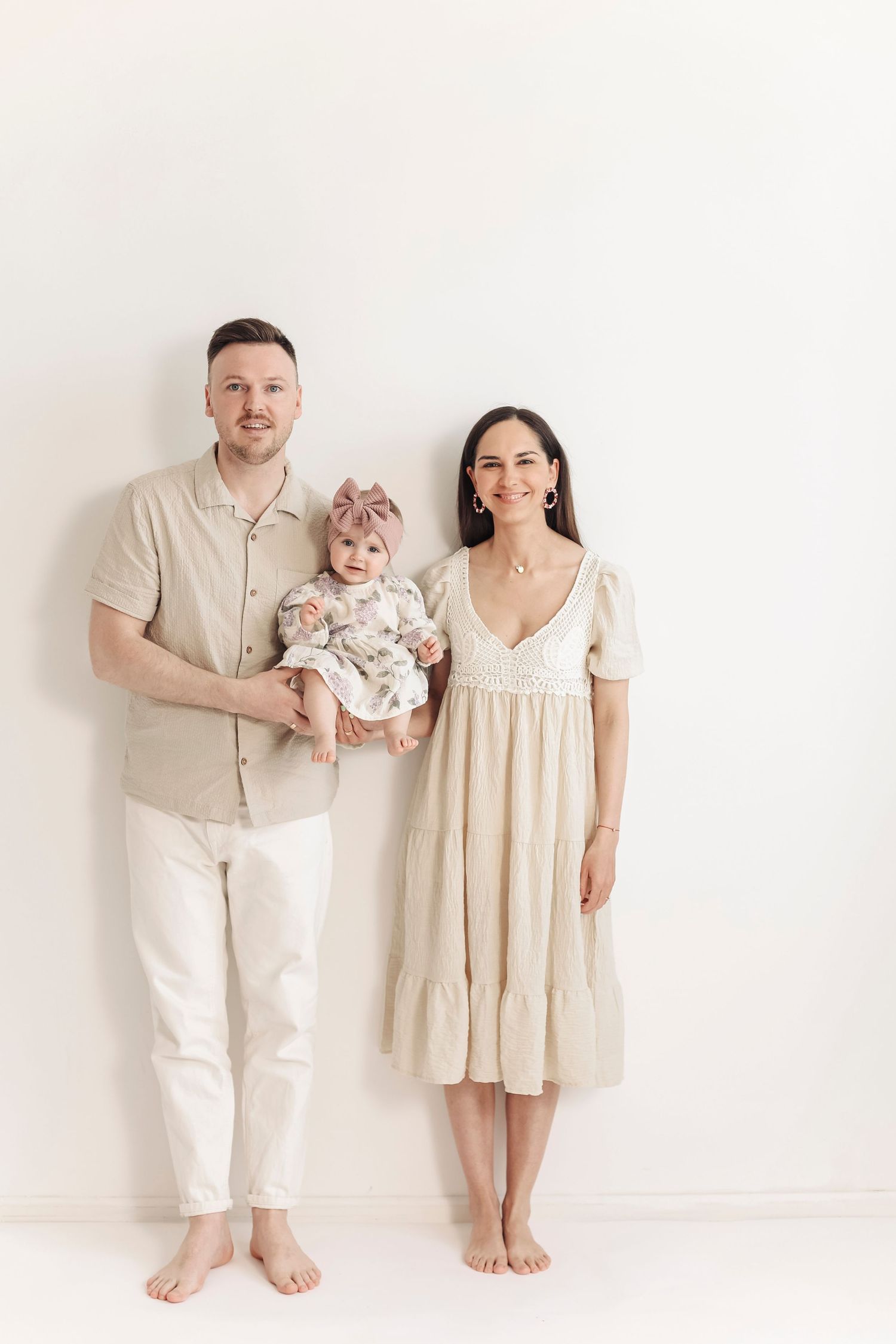 A young family in matching beige outfits poses barefoot against a white wall, parents holding their baby girl.