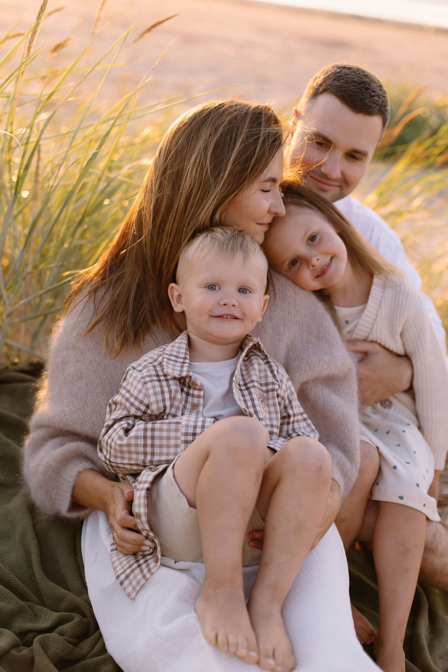 Happy family of four sitting together on a sandy beach at golden hour, parents embracing young children outdoors.