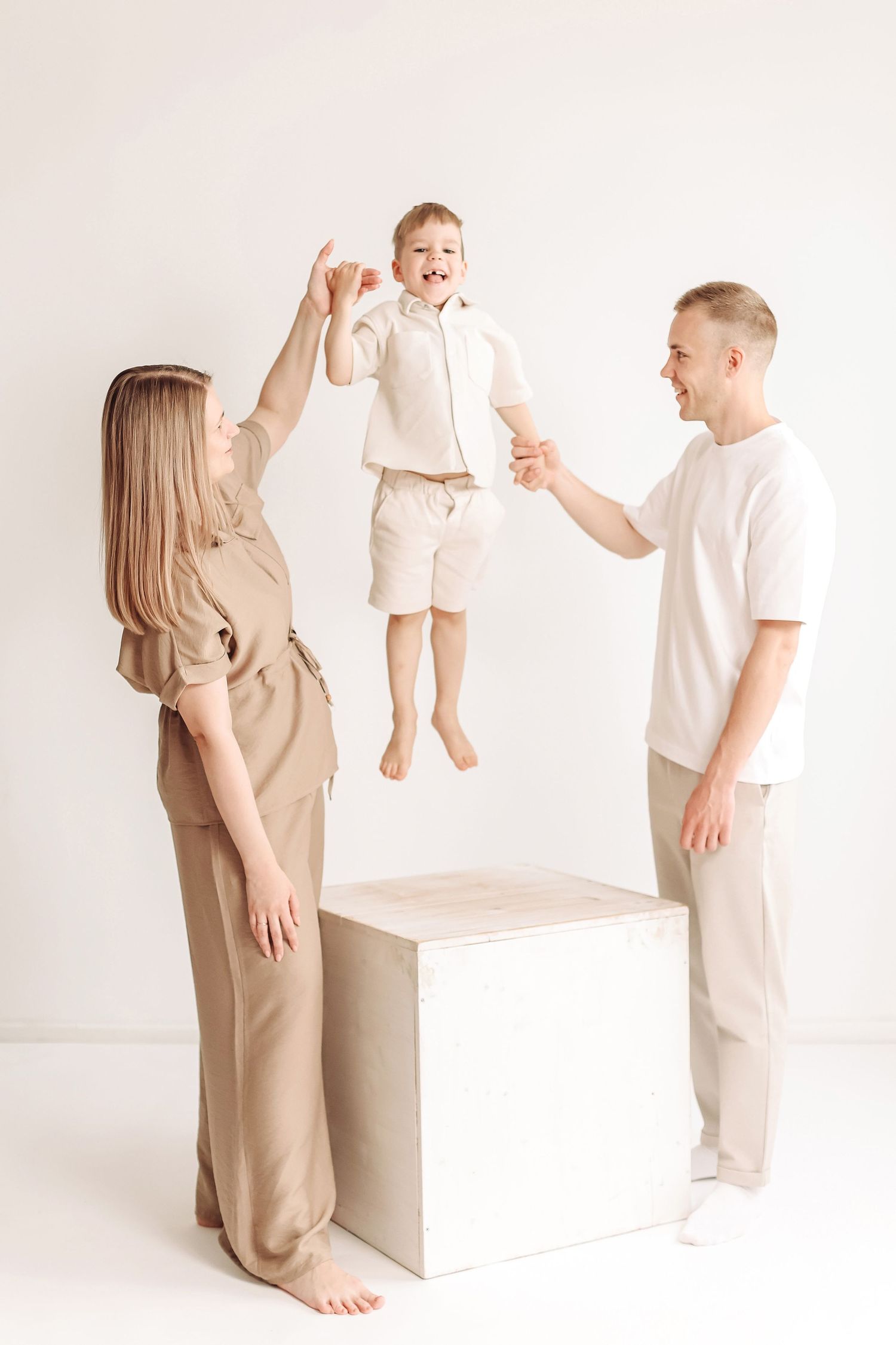 Happy family in beige outfits lifting joyful young boy in bright white studio setting.