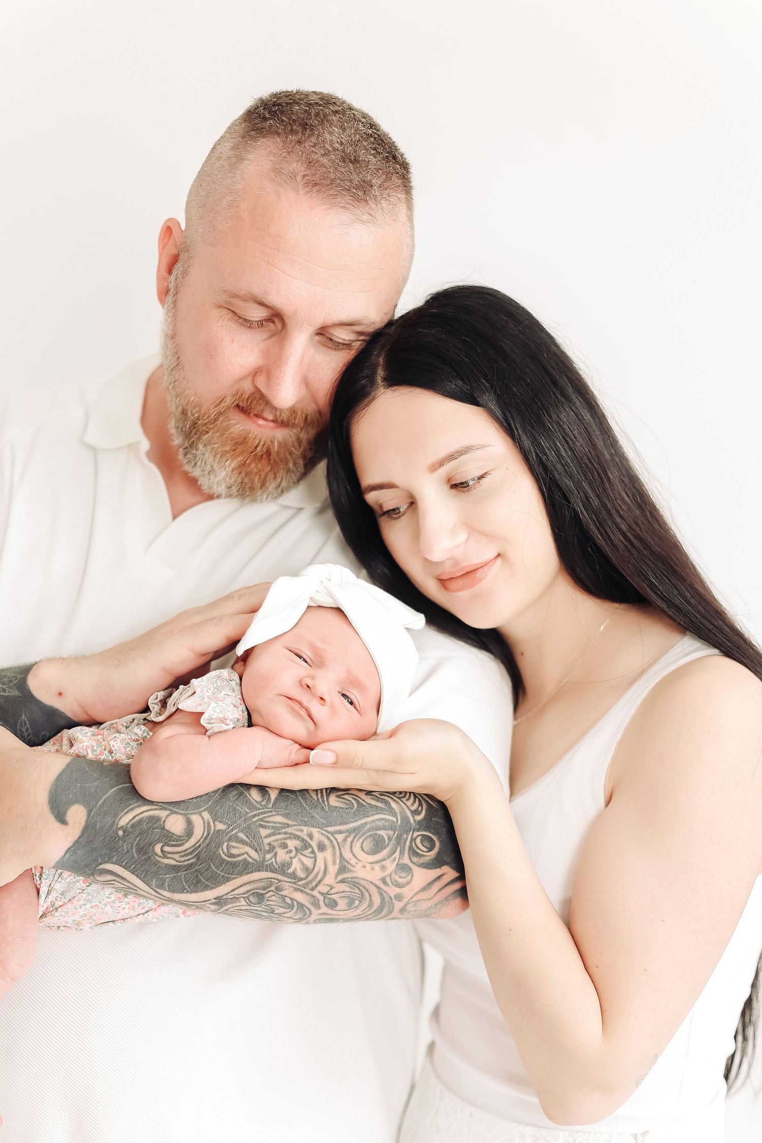 A couple tenderly holds their sleeping newborn baby, the father showing tattoos, against a white background.