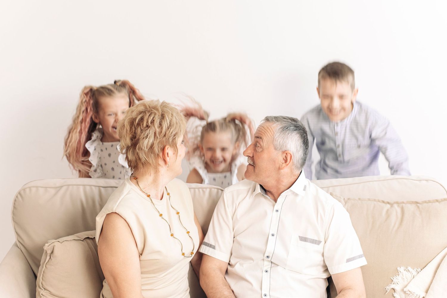 Grandparents sitting on a beige sofa smiling at each other while three playful grandchildren play behind them.