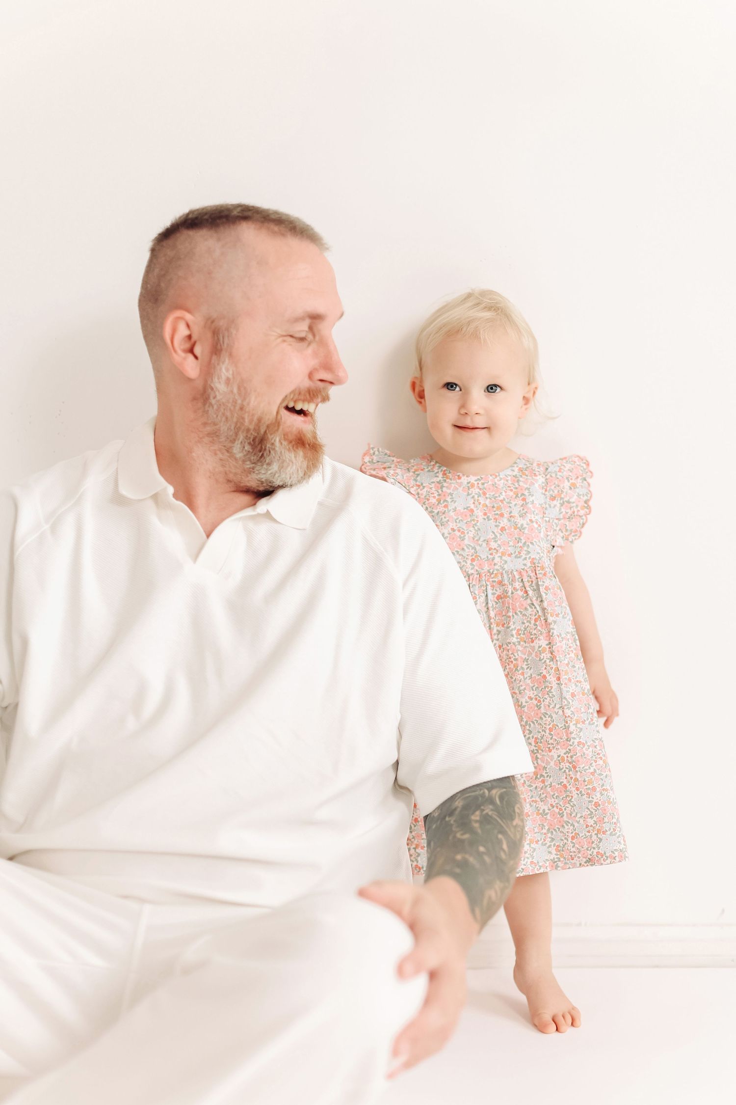 A laughing father in white sits beside his smiling toddler daughter in a floral dress against a white background.