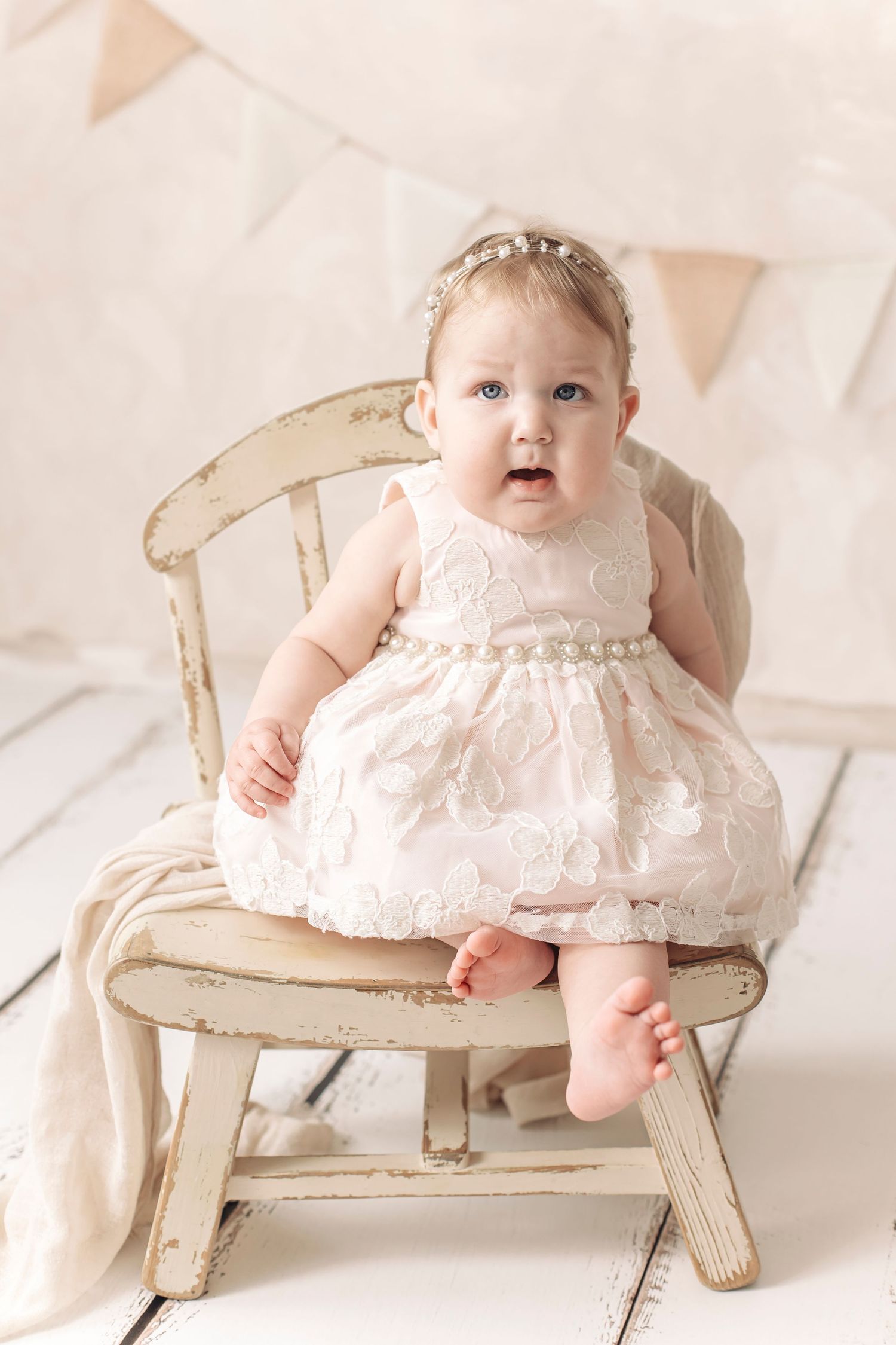 Baby girl in white lace dress with pearl belt sits on vintage chair with bunting backdrop.