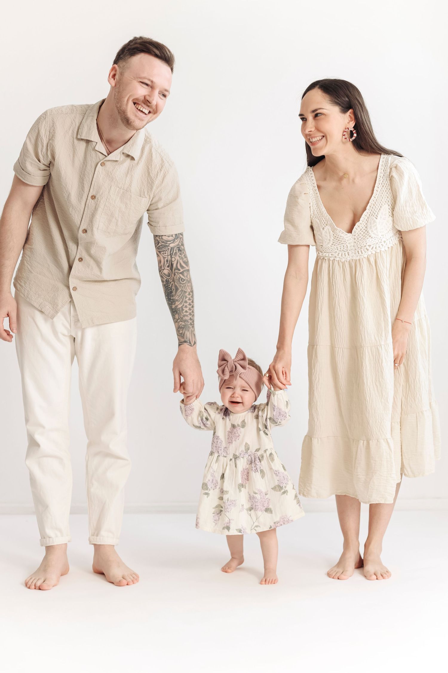 Happy young family with tattooed father, brunette mother in beige dress, and toddler daughter holding hands on white background.