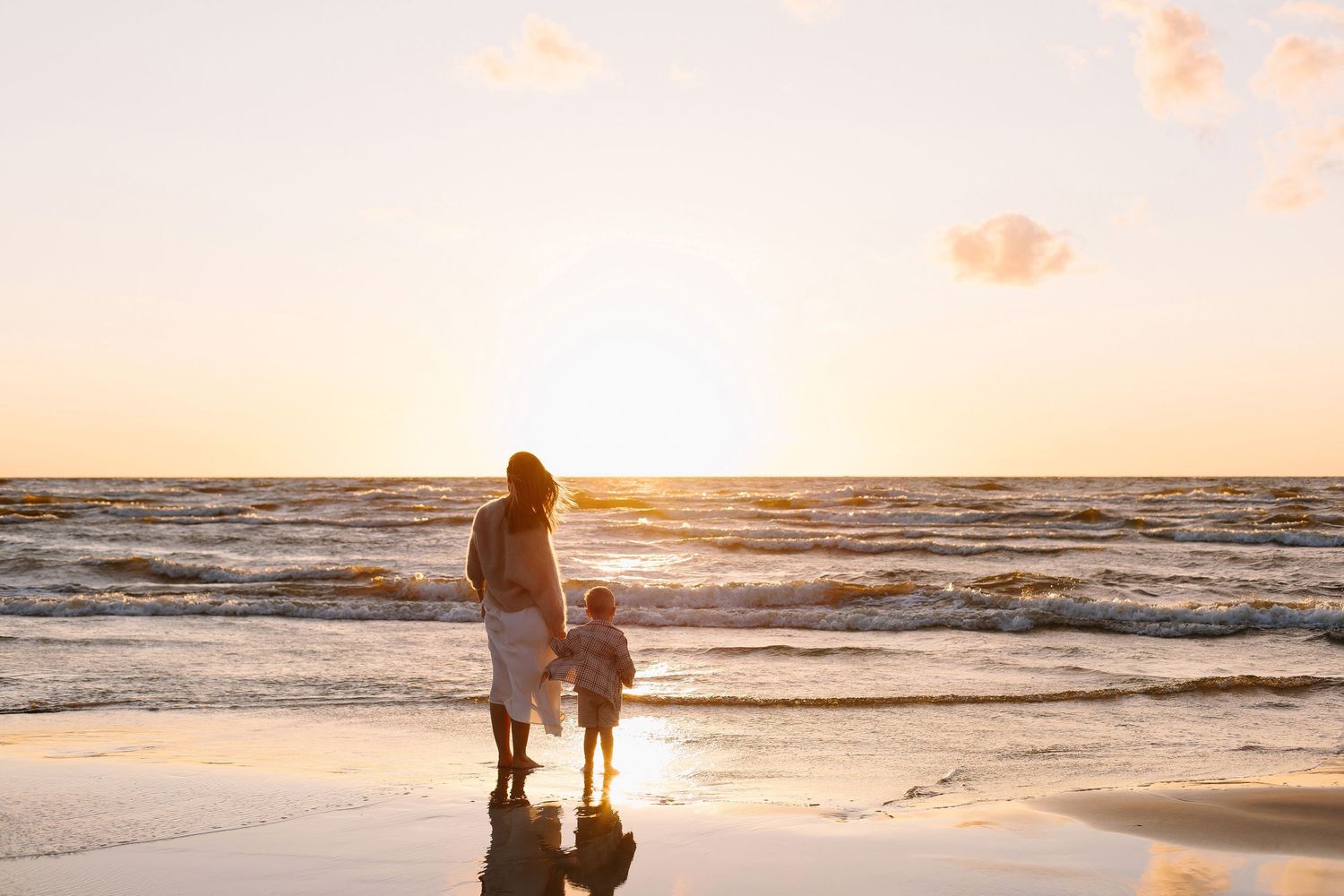 Mother and young child standing together on a beach watching a beautiful golden sunset over the ocean.