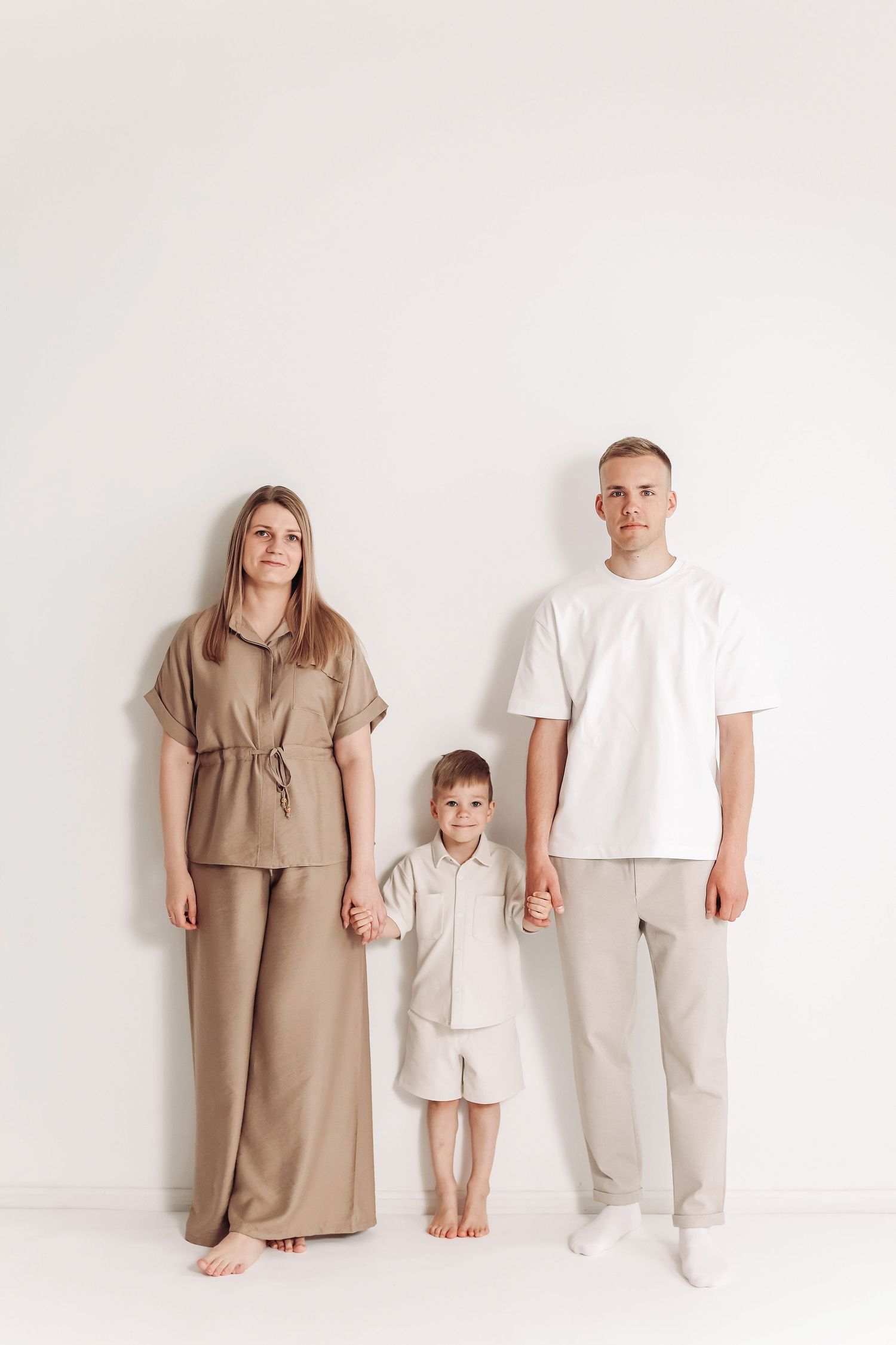 A family of three in neutral beige outfits stand against a white wall, parents holding hands with their young child.