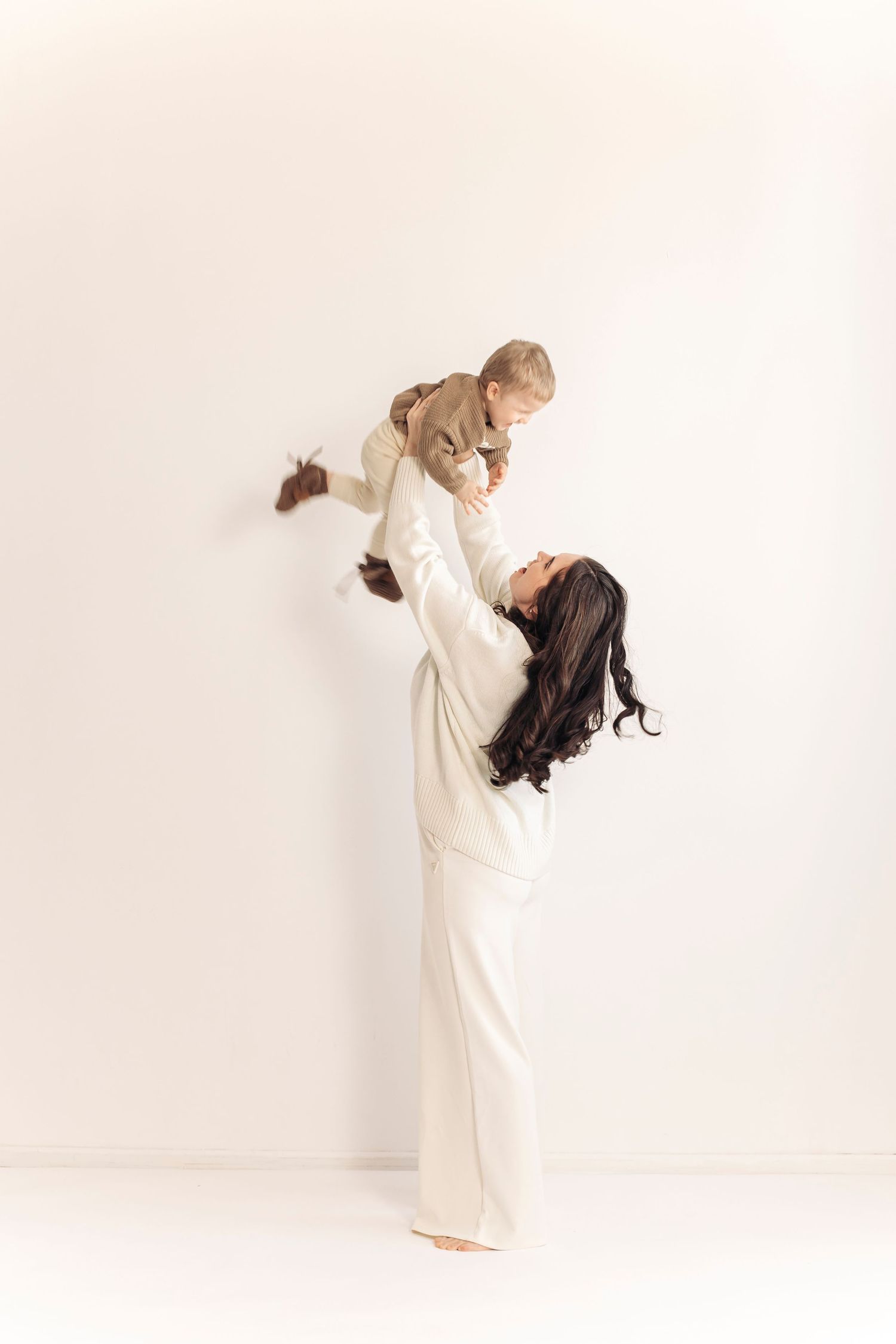 A woman in white outfit joyfully lifts her baby above her head against a clean white background.
