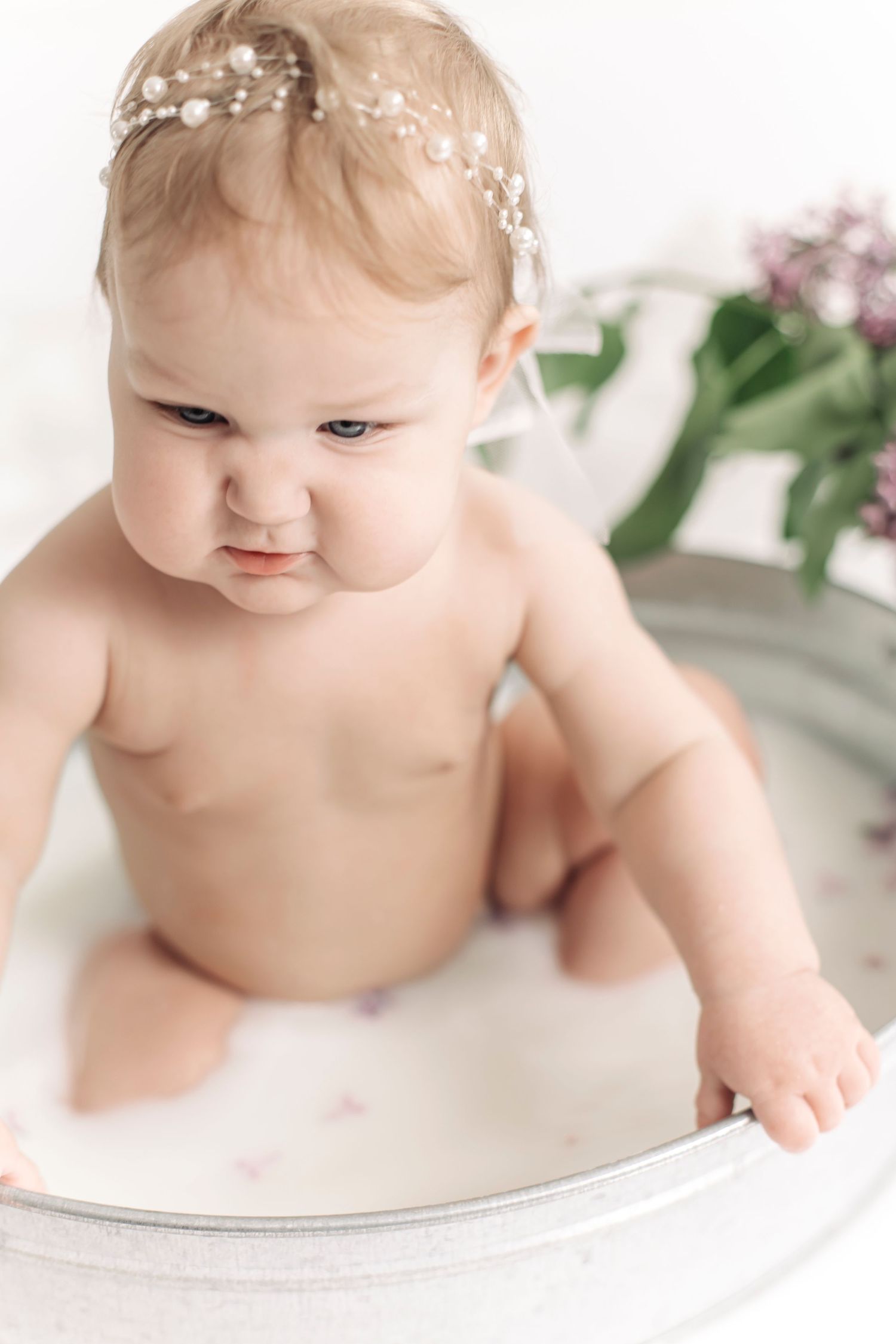 Cute baby girl with pearl headband sitting in milk bath tub with flowers.