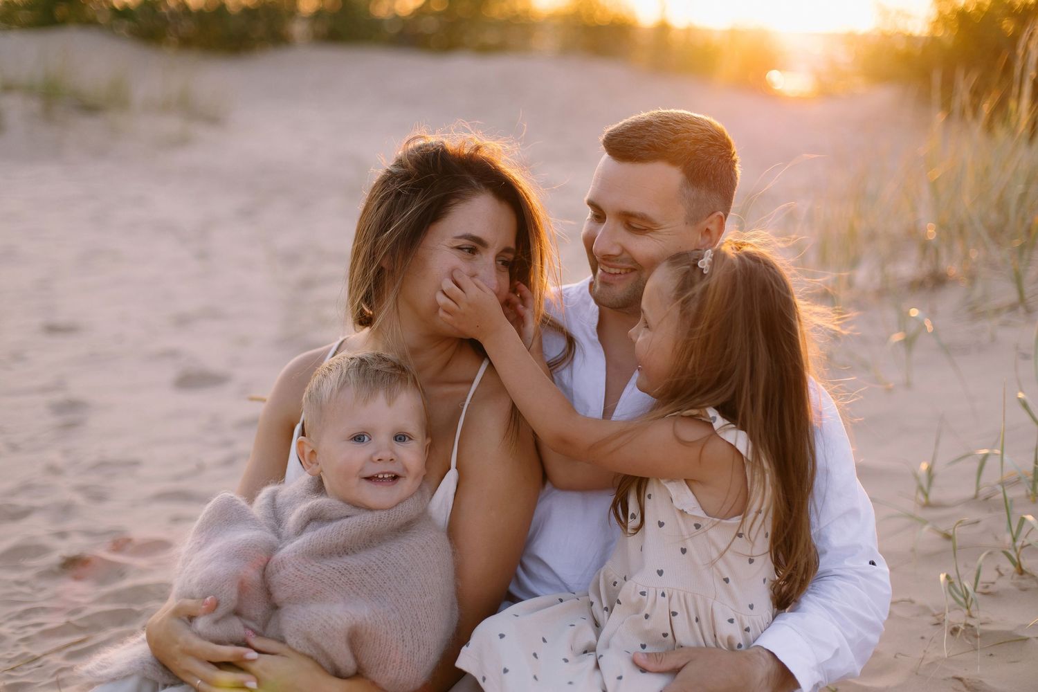 Family of four enjoying a golden sunset beach moment, with parents and two young children sharing a joyful, loving embrace.