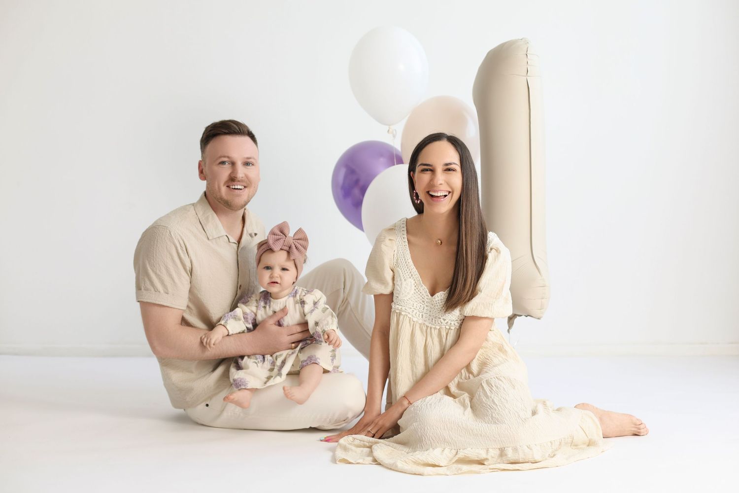 Family photo with parents and baby girl celebrating first birthday with balloons on white background.