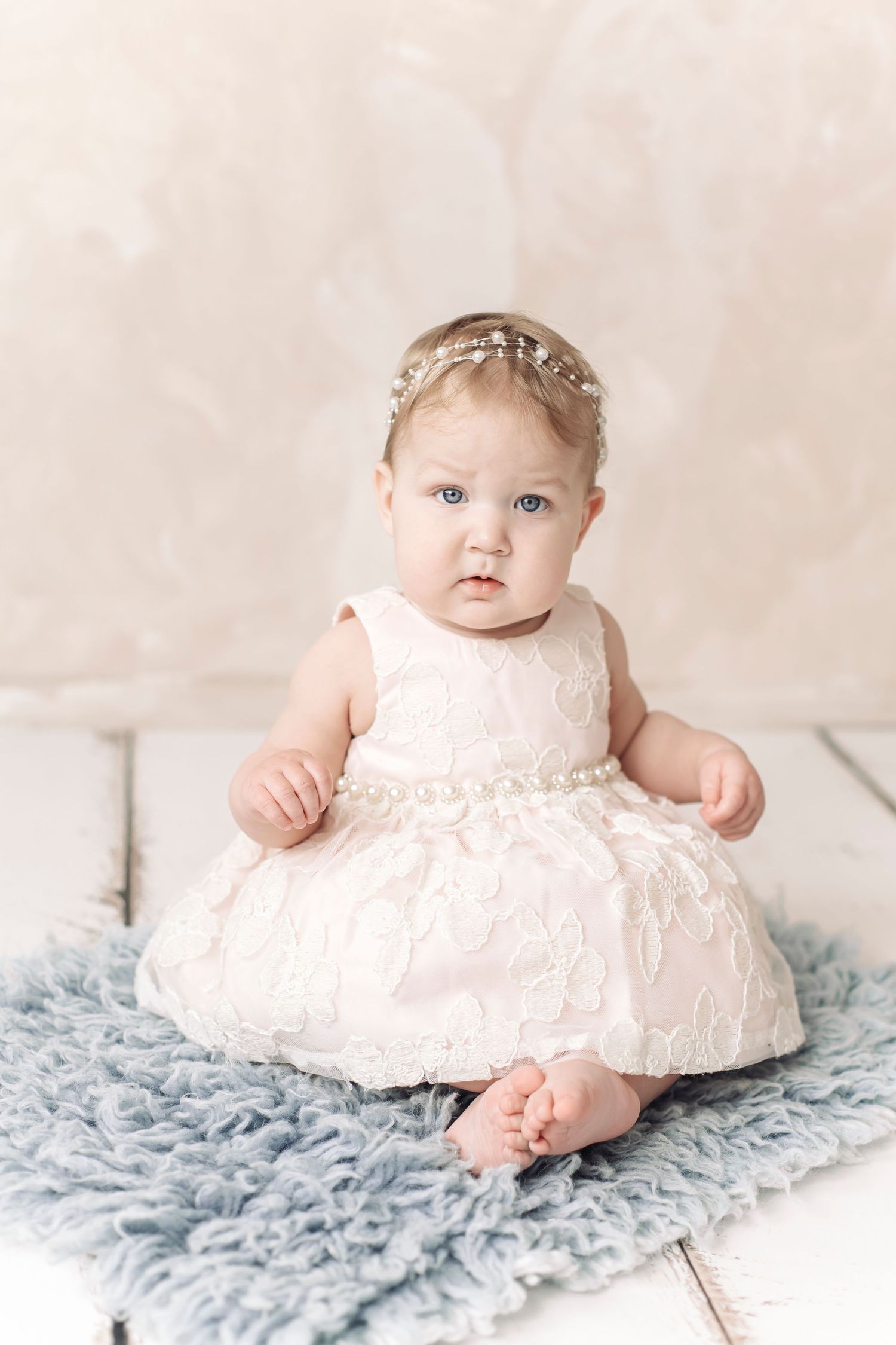 Baby girl in white lace dress and pearl headband sits on blue fur rug for professional portrait photo.