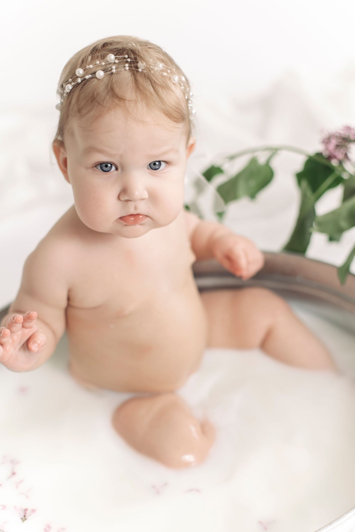 Cute baby girl with pearl headband sitting in a milk bath with flowers and greenery.