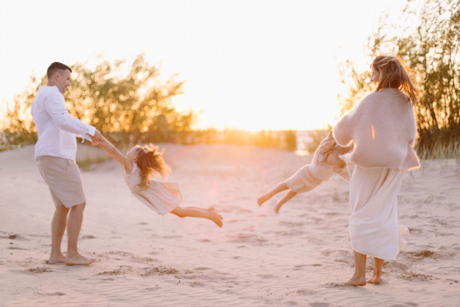 Family playing on sandy beach at golden sunset, parents swinging children in the air with trees in background.