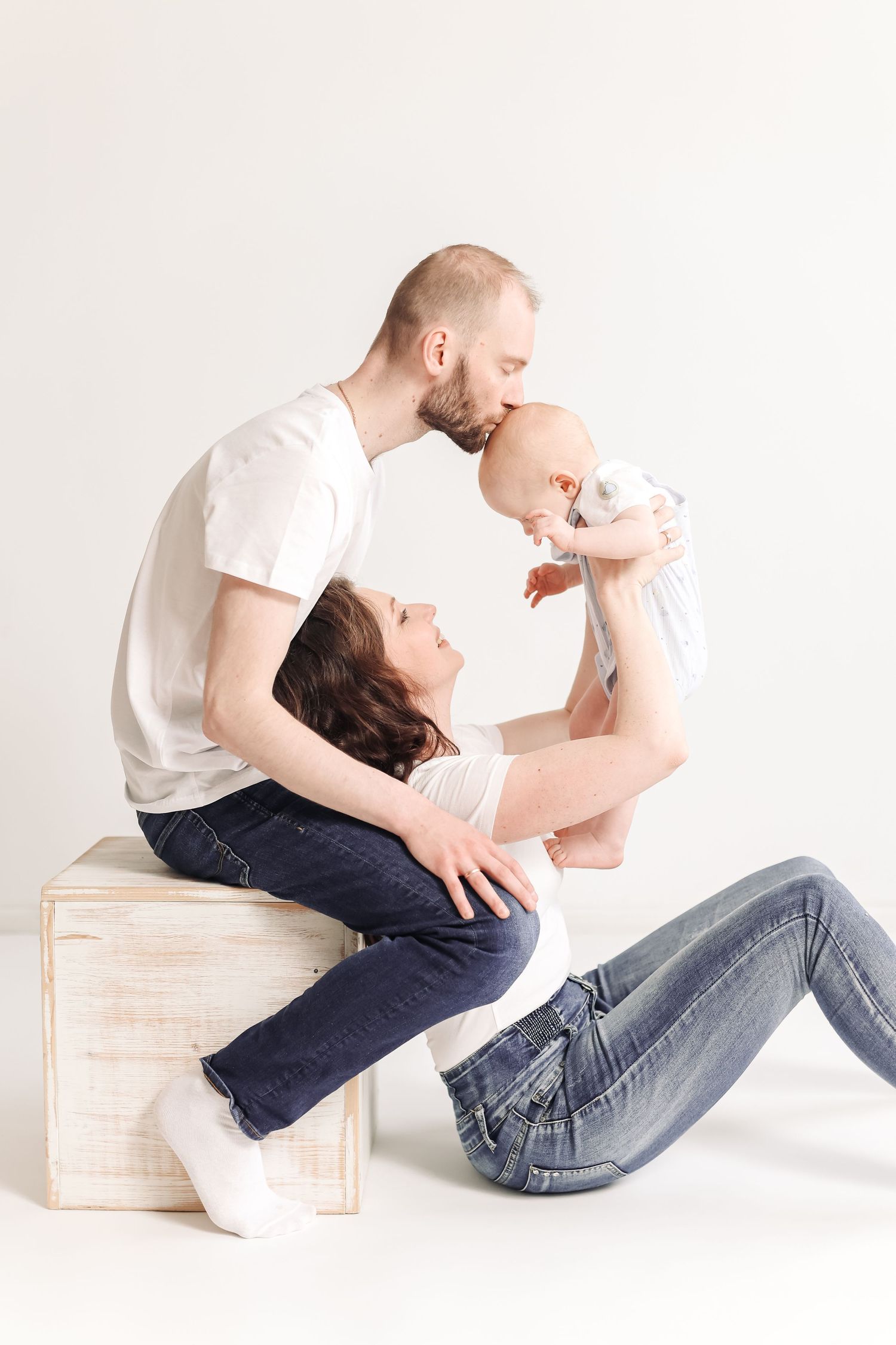 Parents in white shirts and jeans kissing their baby, father seated on wooden cube while mother reclines on white floor.