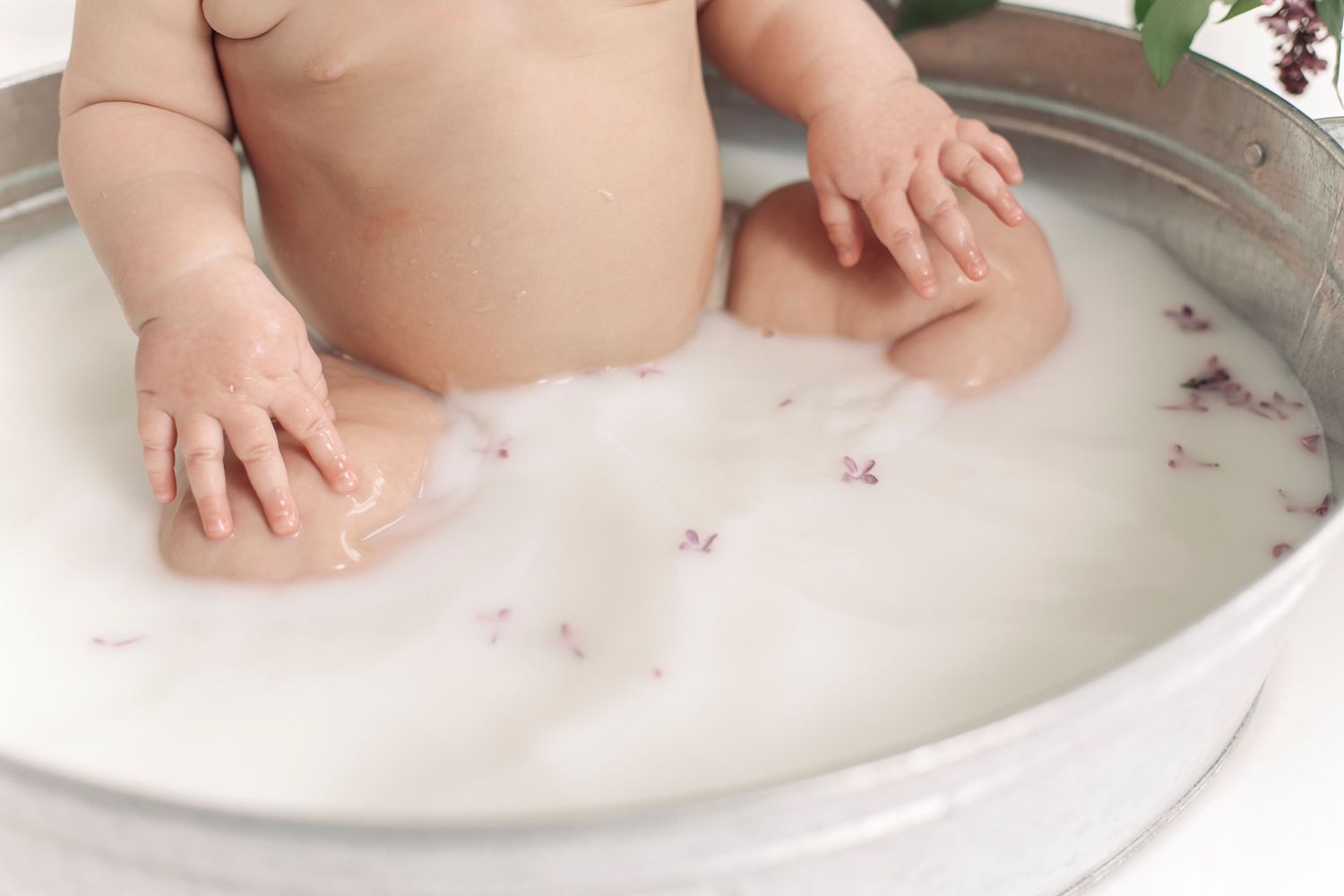 Baby sitting in a round metal tub filled with white milk bath and scattered flower petals.