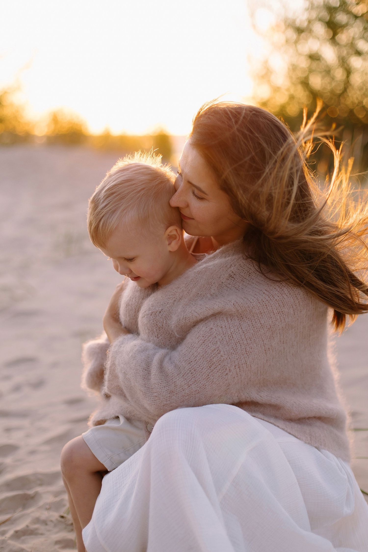 Loving mother gently kissing her toddler while sitting on sandy beach during warm golden sunset.