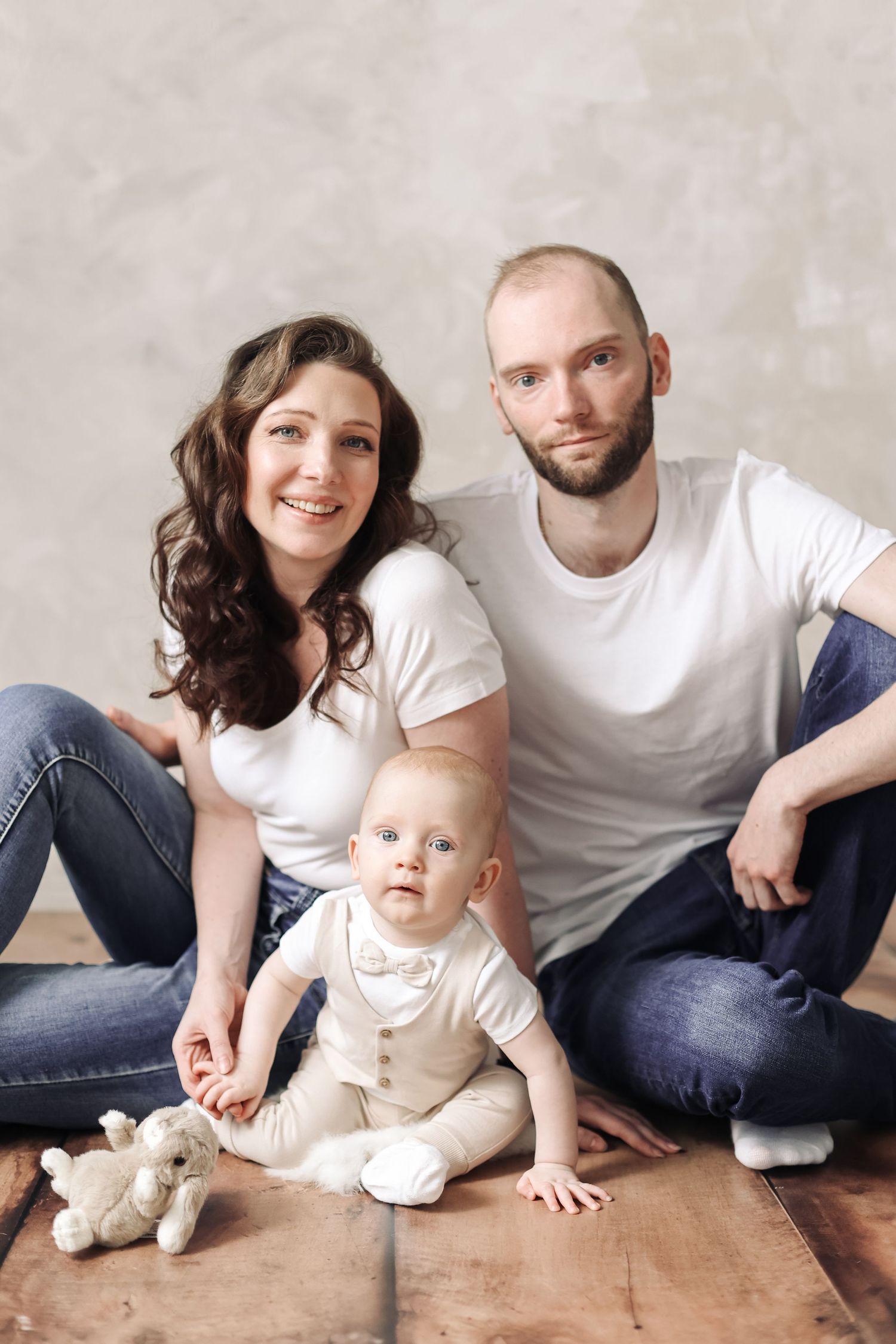 Young family with baby sitting on wooden floor, parents in white tees and jeans, infant in bow tie outfit with stuffed toy.