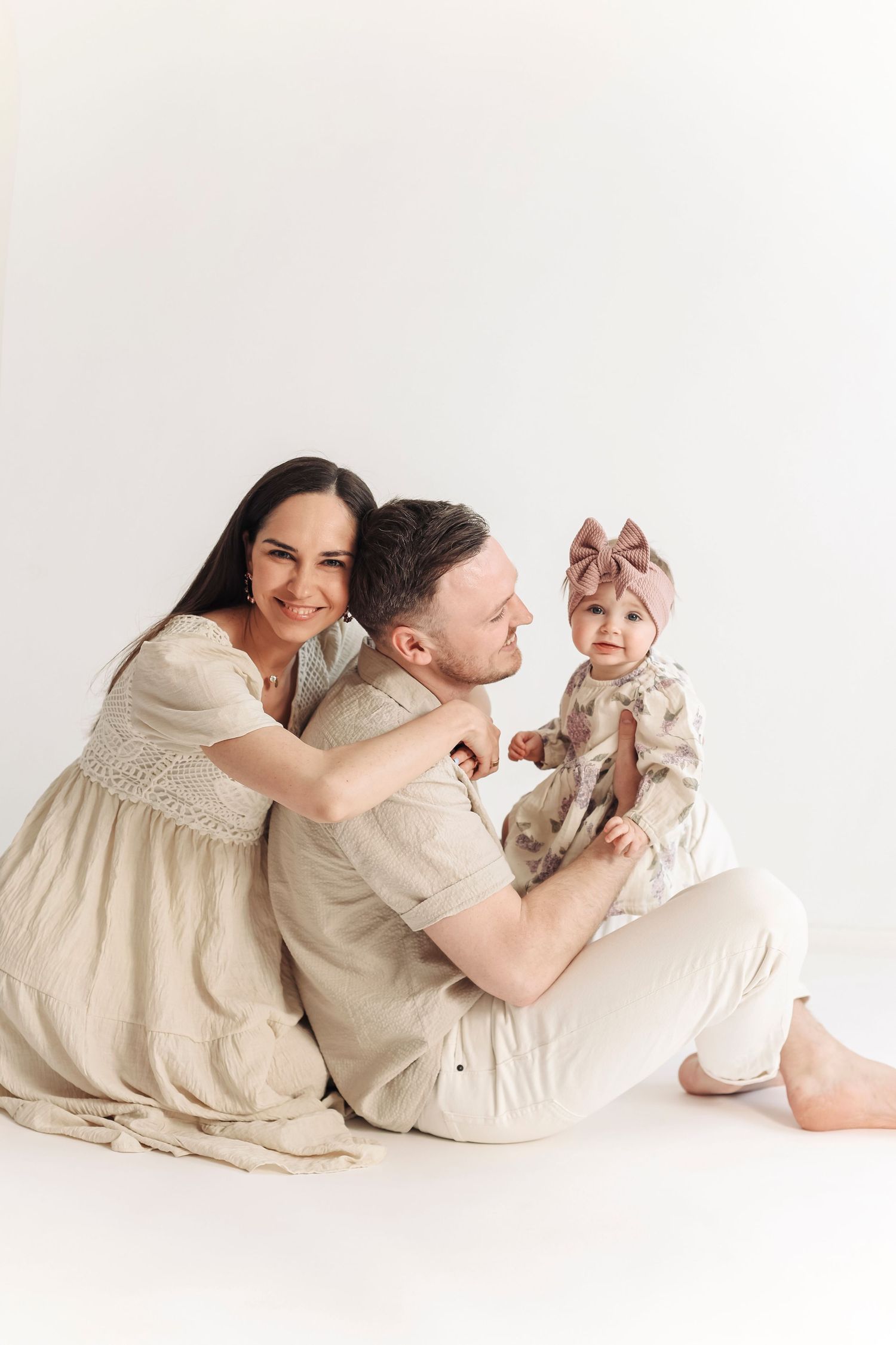 A happy young family with a mother, father, and baby girl in neutral tones sitting together on a white background.