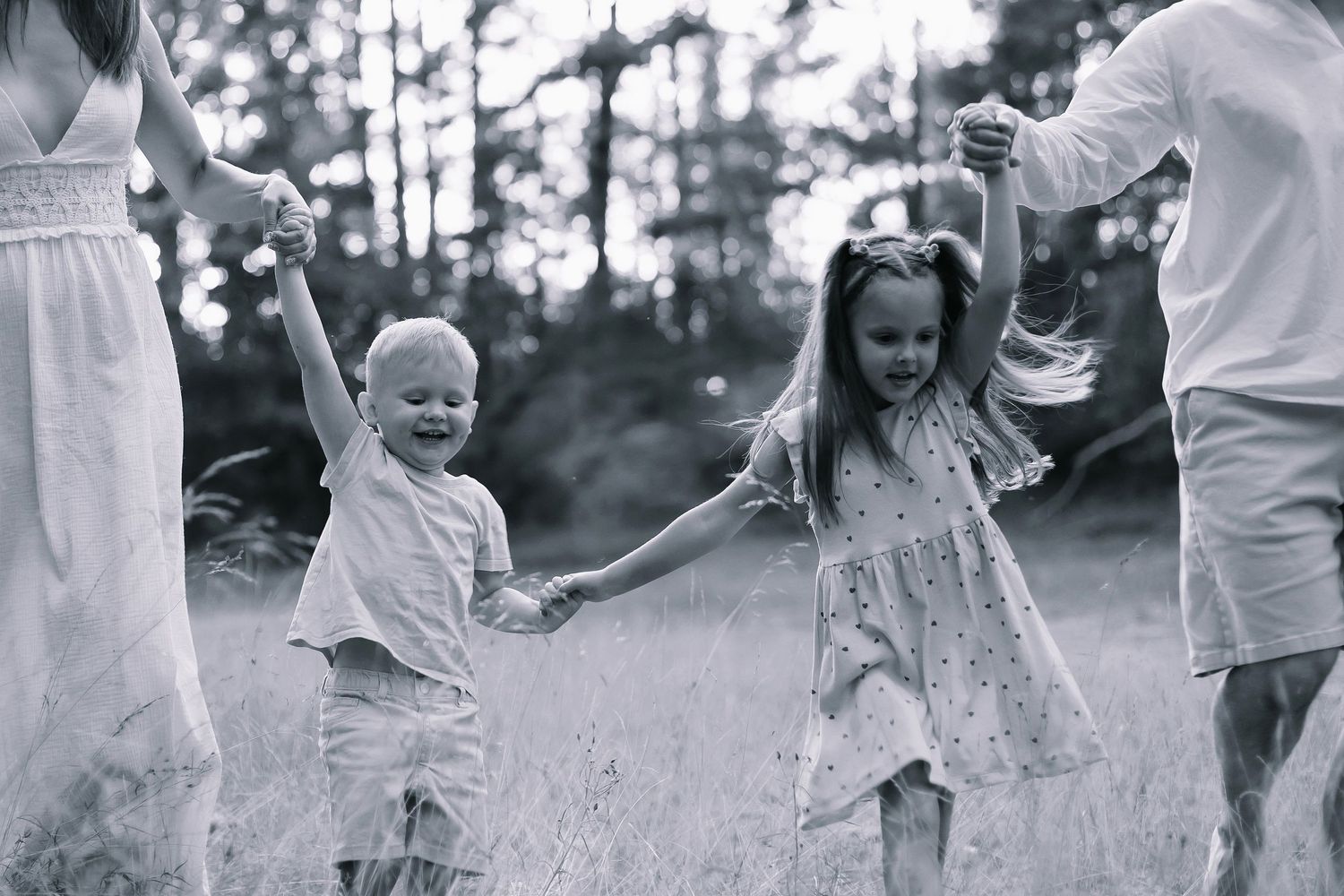 Black and white photo of two young children joyfully holding hands with parents in a sunlit meadow.