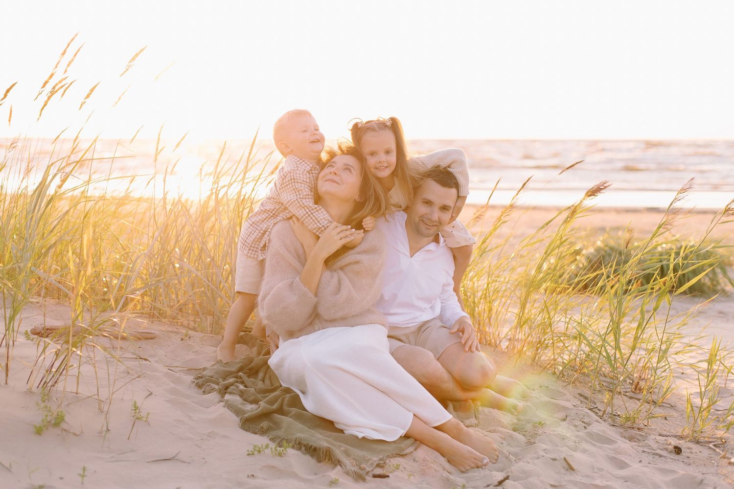 Happy family of four embracing on sandy beach dunes in warm golden sunlight.