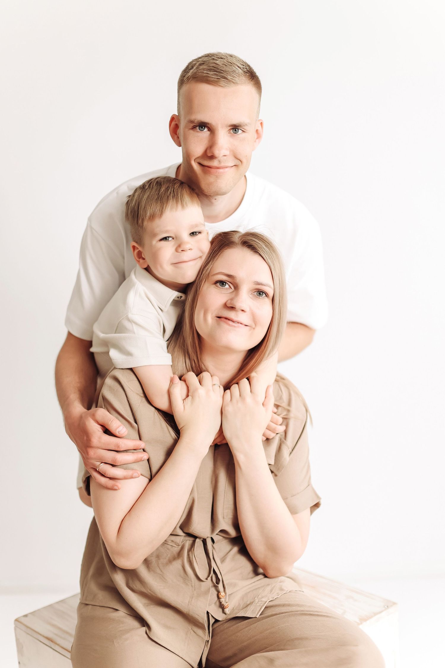 A young family of three posing together, father and toddler son embracing mother in a bright white studio setting.