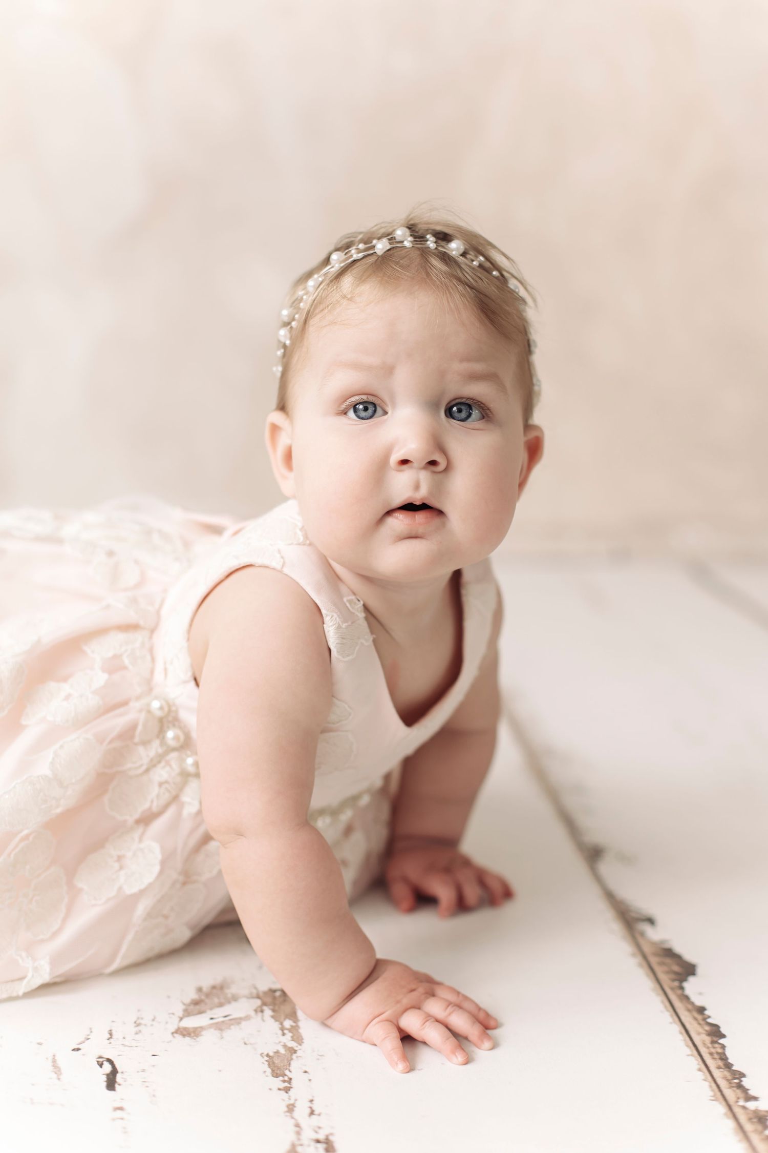 Baby girl in pink lace dress and pearl headband crawling on white wooden floor during professional photo session.
