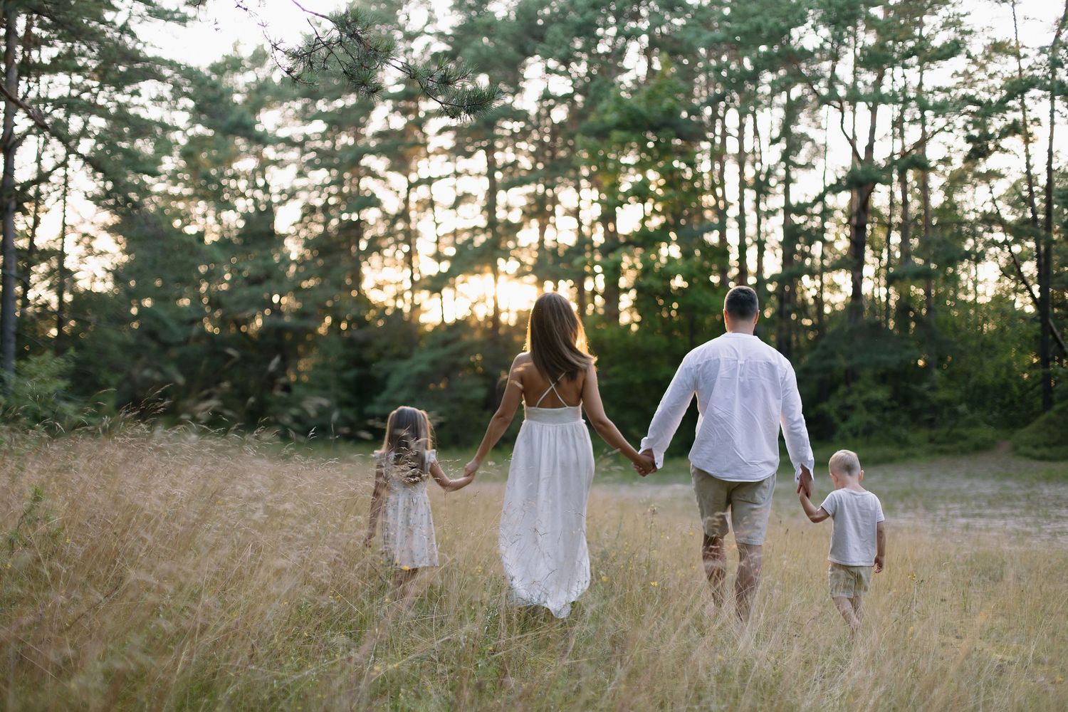 Family of four holding hands walking through golden meadow toward sunset forest in white summer outfits.