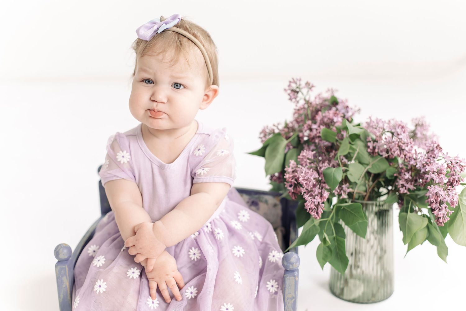 Baby girl in lavender dress with bow headband sits beside purple lilac flowers in vintage vase on white background.