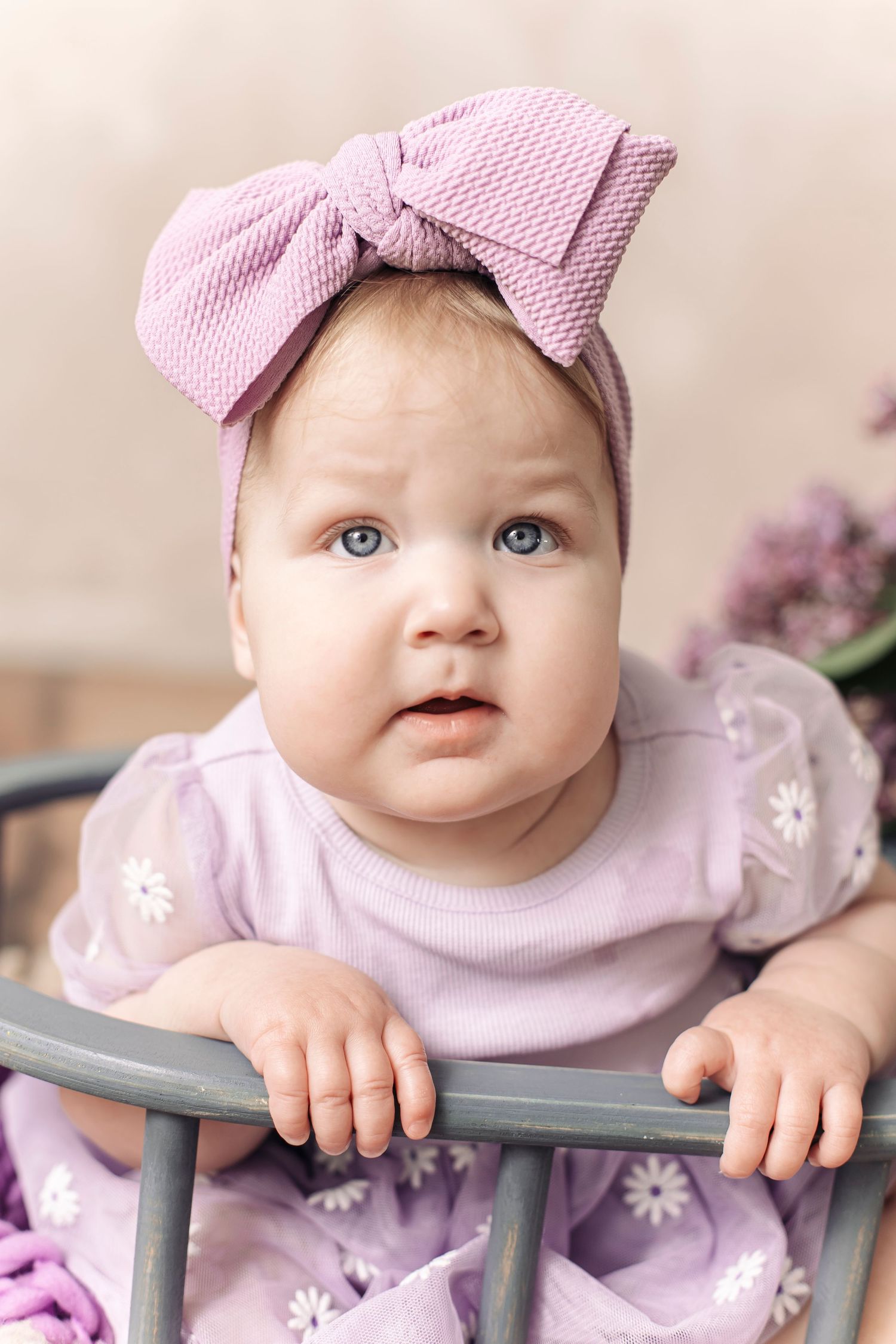 Adorable baby girl in pink floral dress and bow headband standing in a crib with blue eyes.